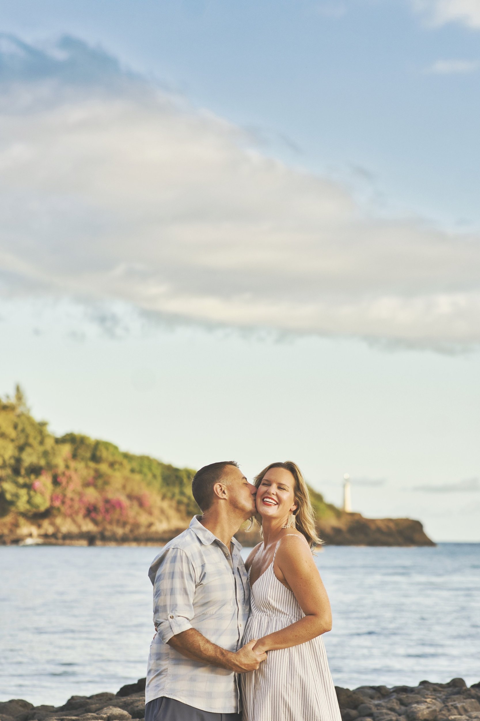 Couple sharing a kiss on a rocky Kauai shoreline with the ocean behind them.