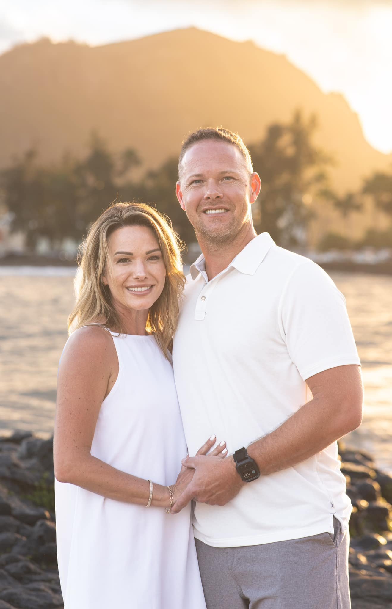 Smiling couple in white posing on a Kauai beach with a golden mountain behind them.