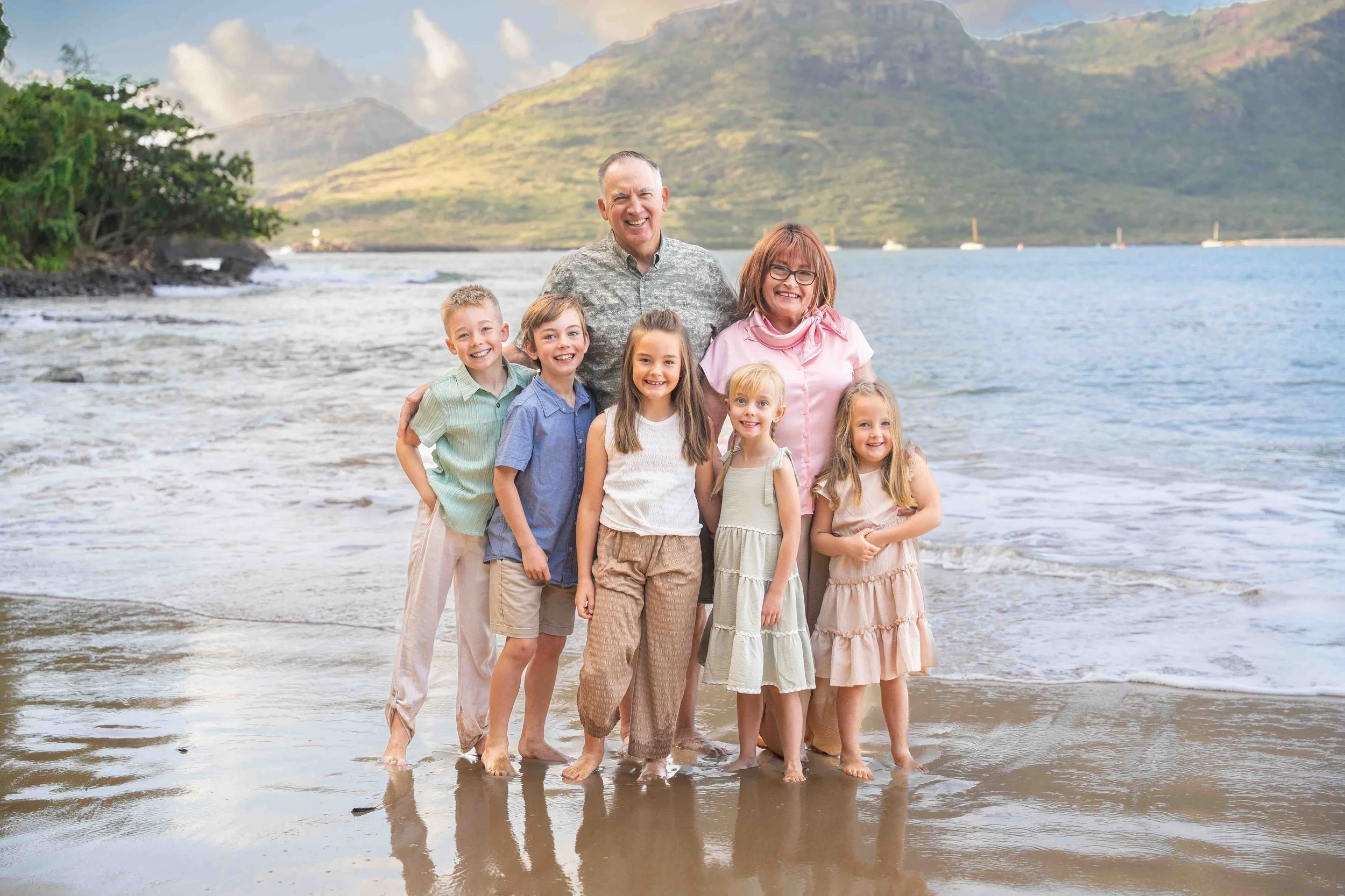 Grandparents pose with their five grandkids on a Kauai beach with waves and mountains behind them.
