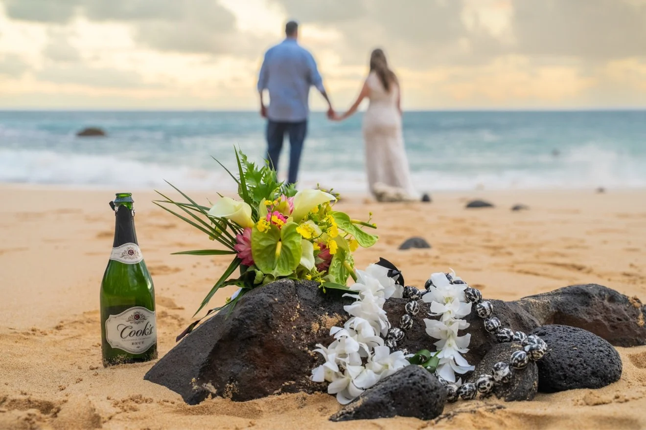 Wedding bouquet, leis and a bottle of champagne resting on lava rocks on a Kauai beach, with the bride and groom holding hands out of focus by the ocean behind.