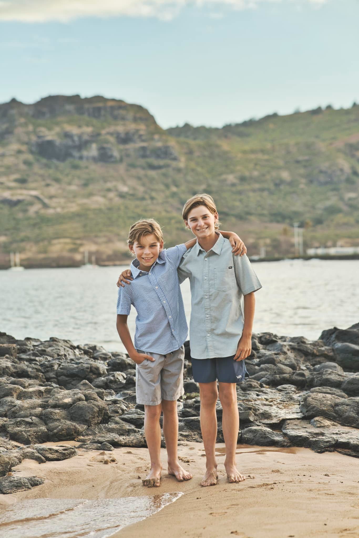 Two brothers with arms around each other smiling on a rocky Kauai beach.
