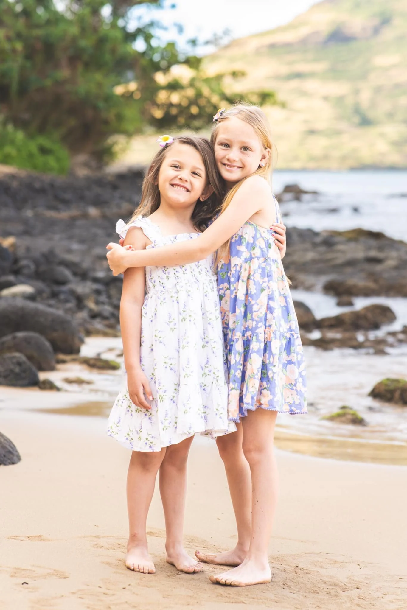 Two sisters in floral dresses hugging and smiling on a sandy Kauai beach.