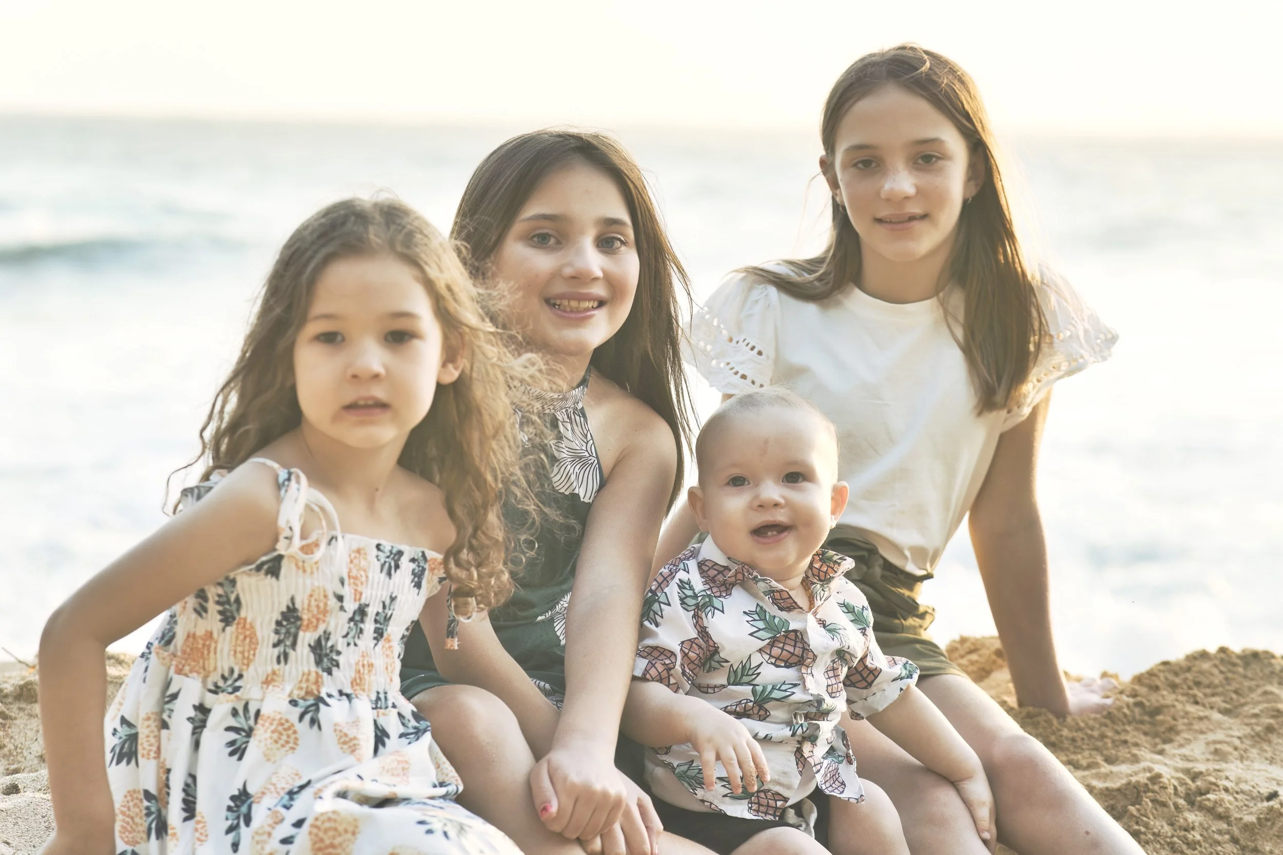 Four kids sitting together in the sand on a Kauai beach with the ocean behind them.