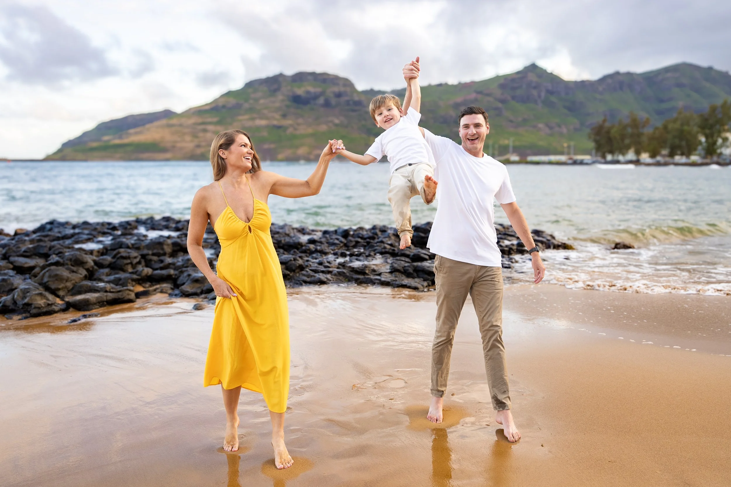 Parents joyfully swinging their young son on a rocky Kauai beach with mountains behind them.
