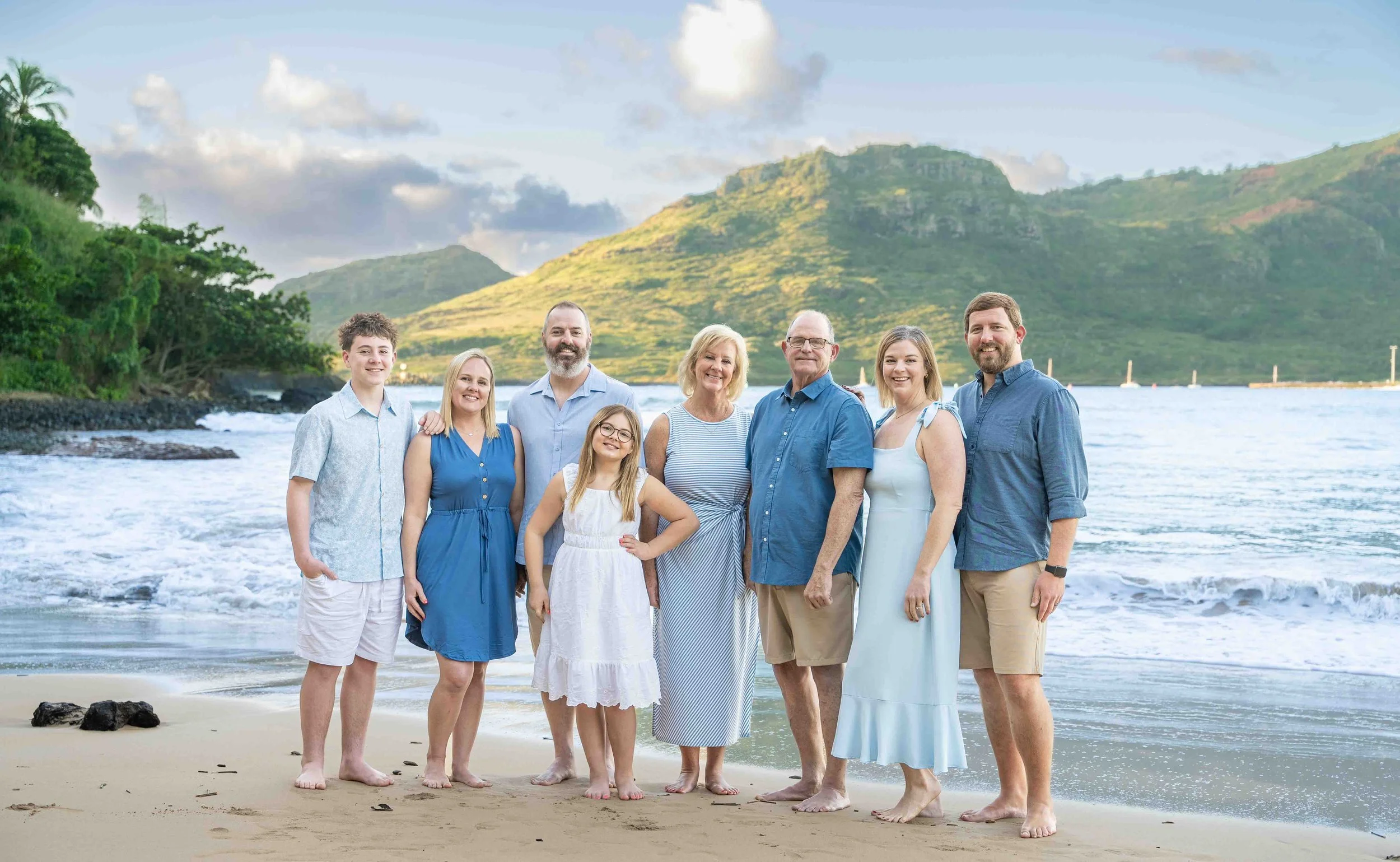 Extended family in coordinated blue and white outfits poses on a Kauai beach beneath green mountains.