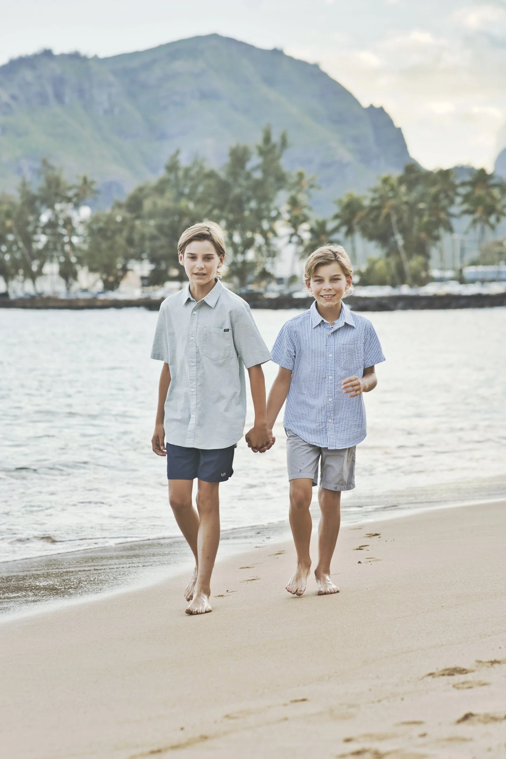Two brothers walking hand in hand toward the camera on a sandy Kauai beach.