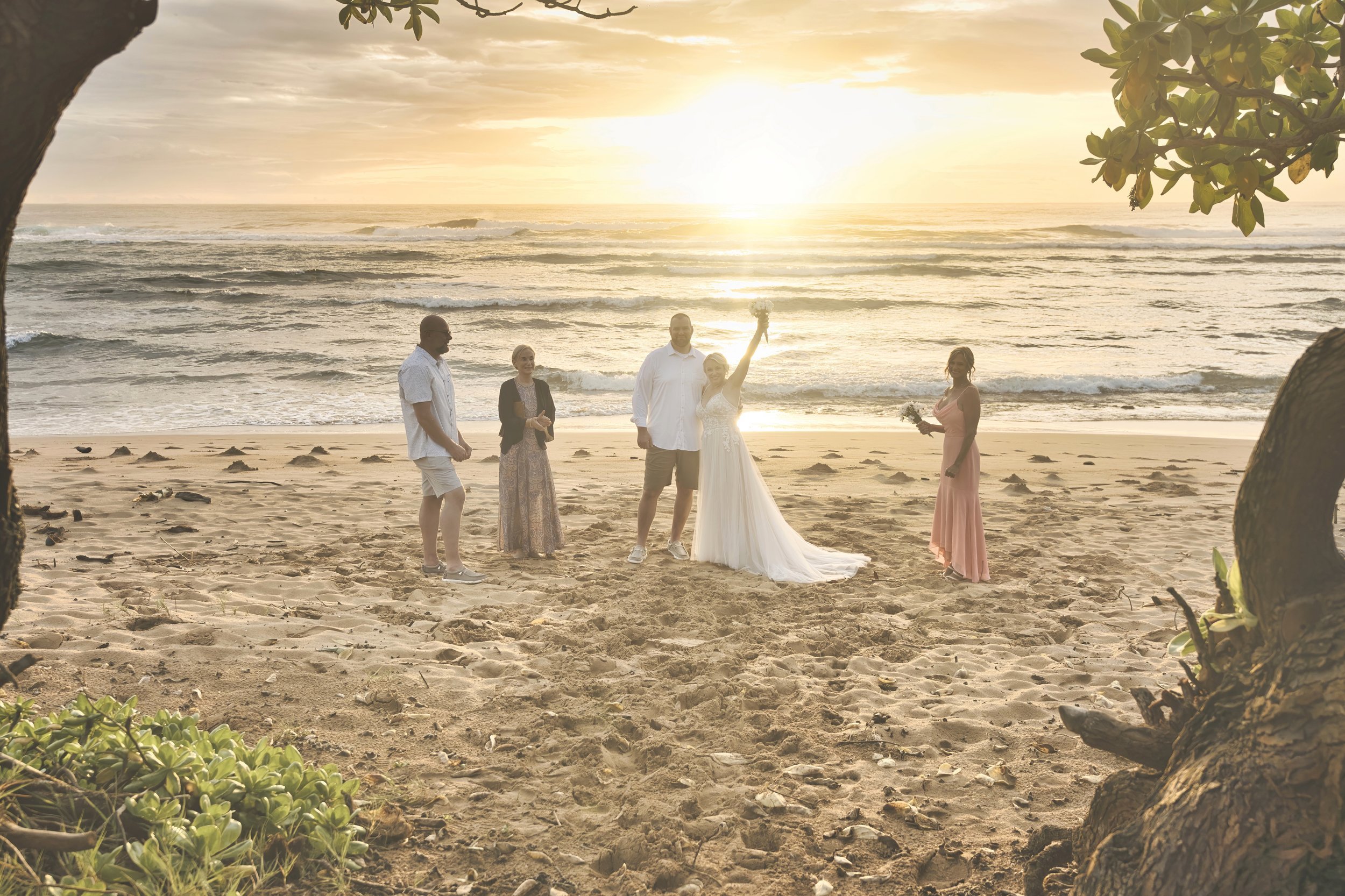 Bride raising her bouquet in celebration during an intimate beach wedding ceremony on Kauai at sunset.