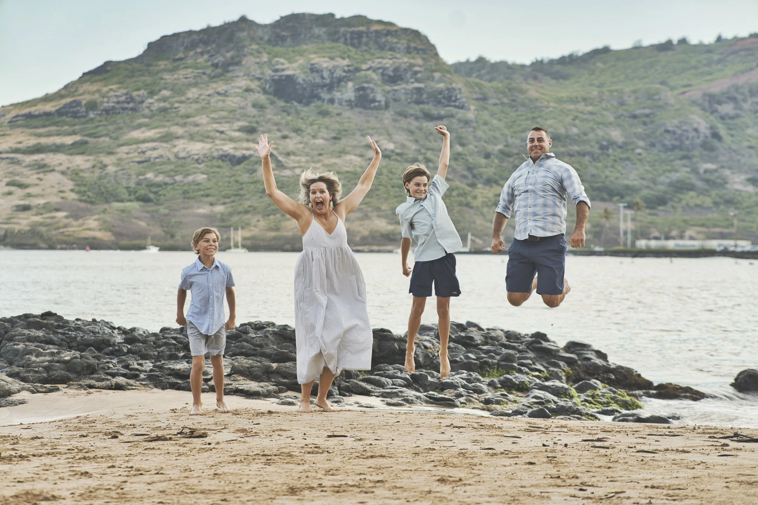 Family of four jumping and laughing on a Kauai beach with green mountains behind.