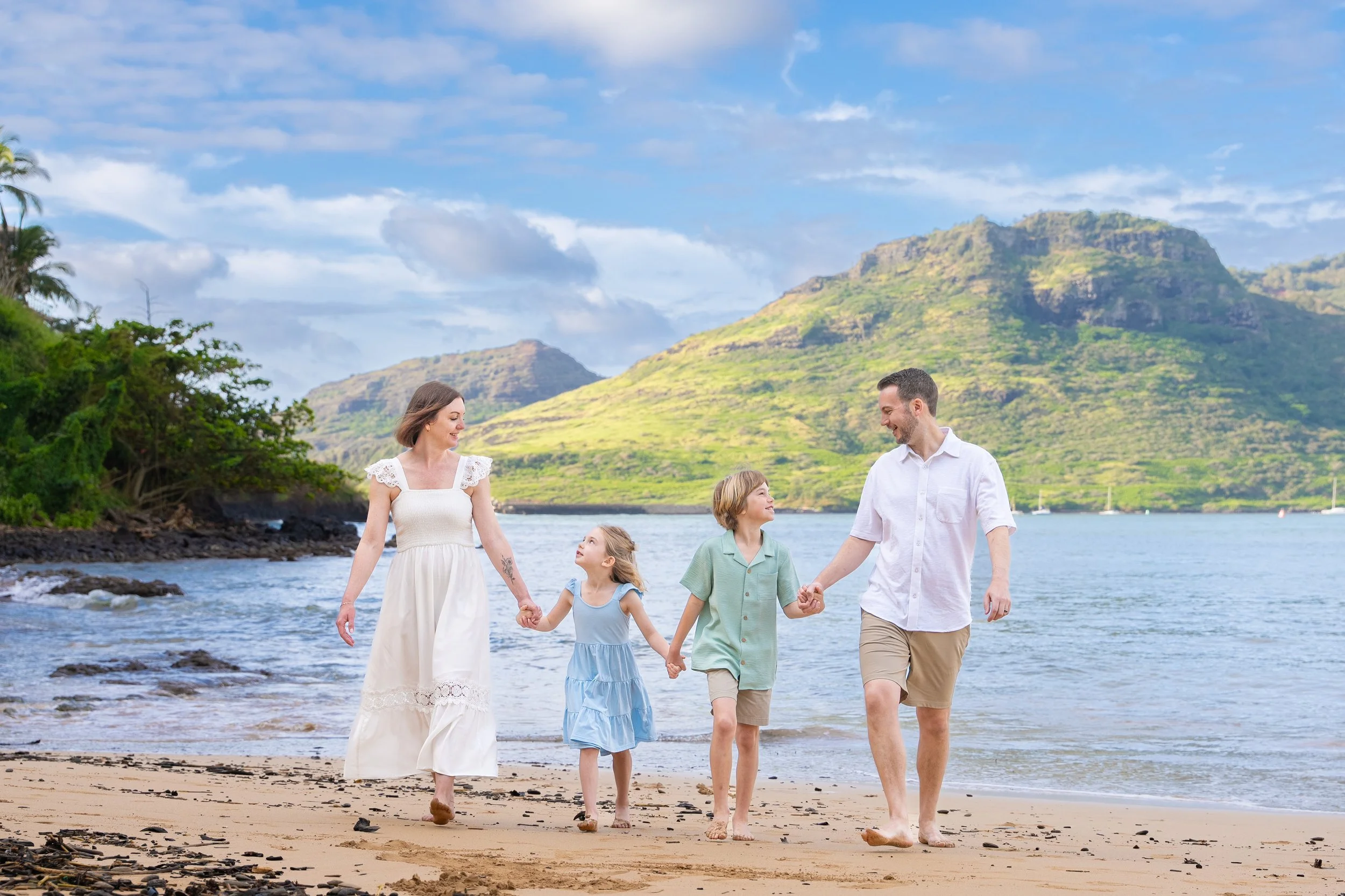 Family of four walking hand in hand along a Kauai beach with green mountains behind them.