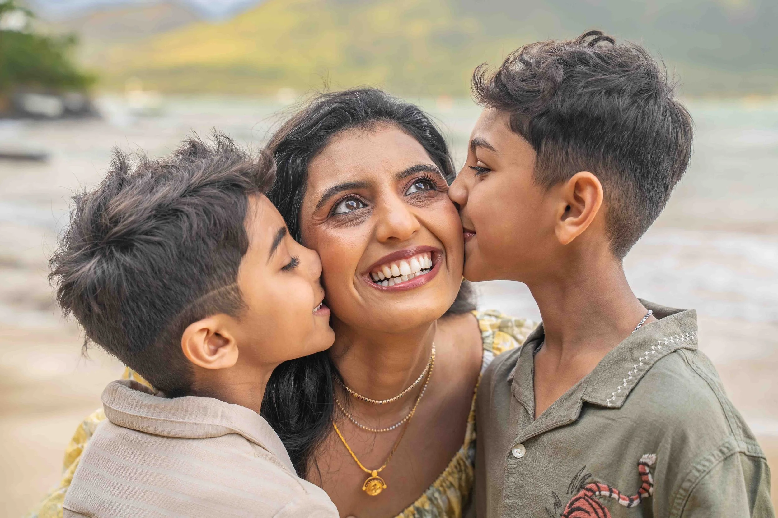 Mom laughing while her two sons kiss her cheeks on a Kauai beach.