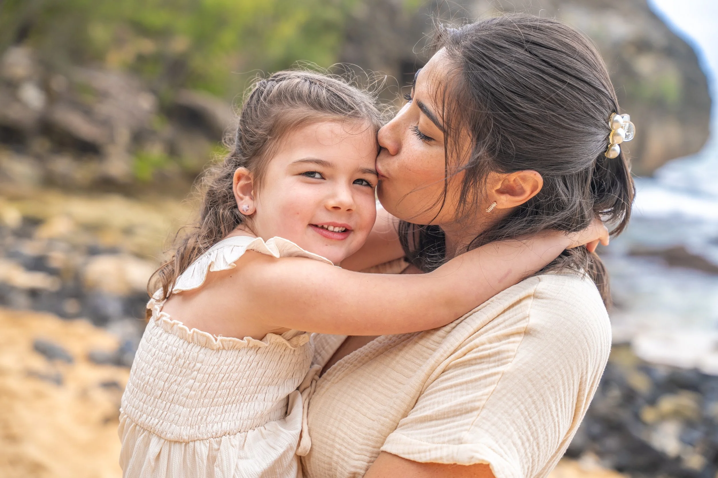 Mom kissing her smiling daughter's temple on a rocky Kauai beach.