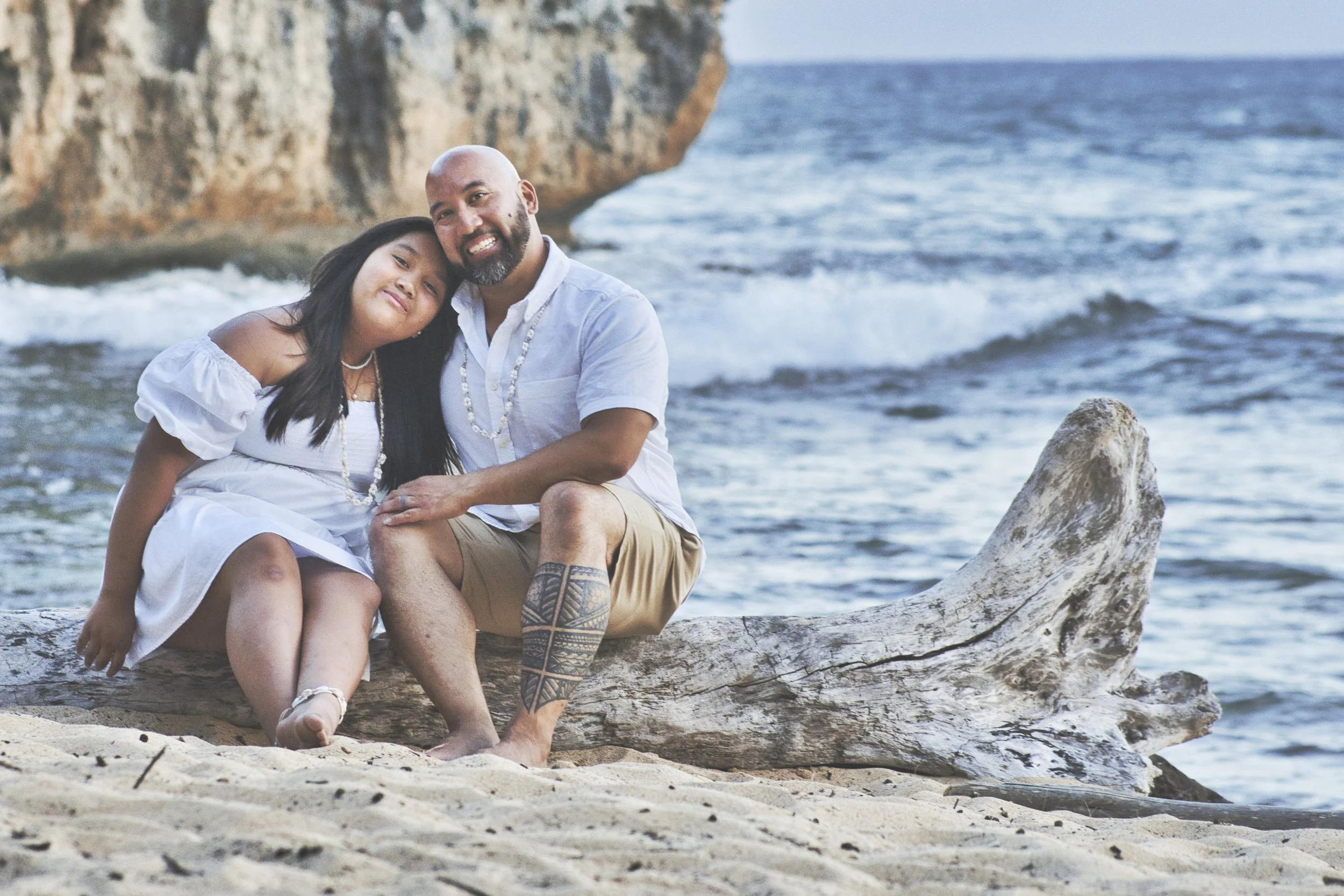 Couple embracing while sitting on driftwood on the beach at Shipwreck Beach, Kauai.