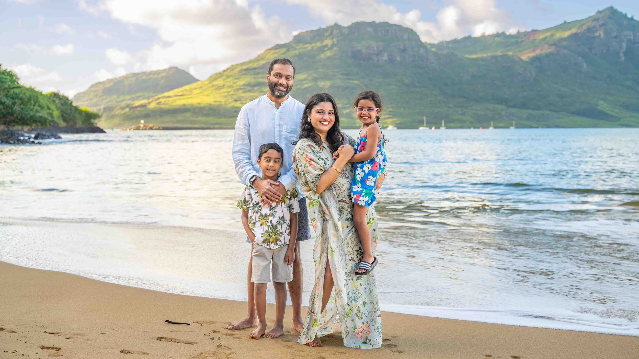 Family of four posing on a Kauai beach with the ocean and mountains behind them.