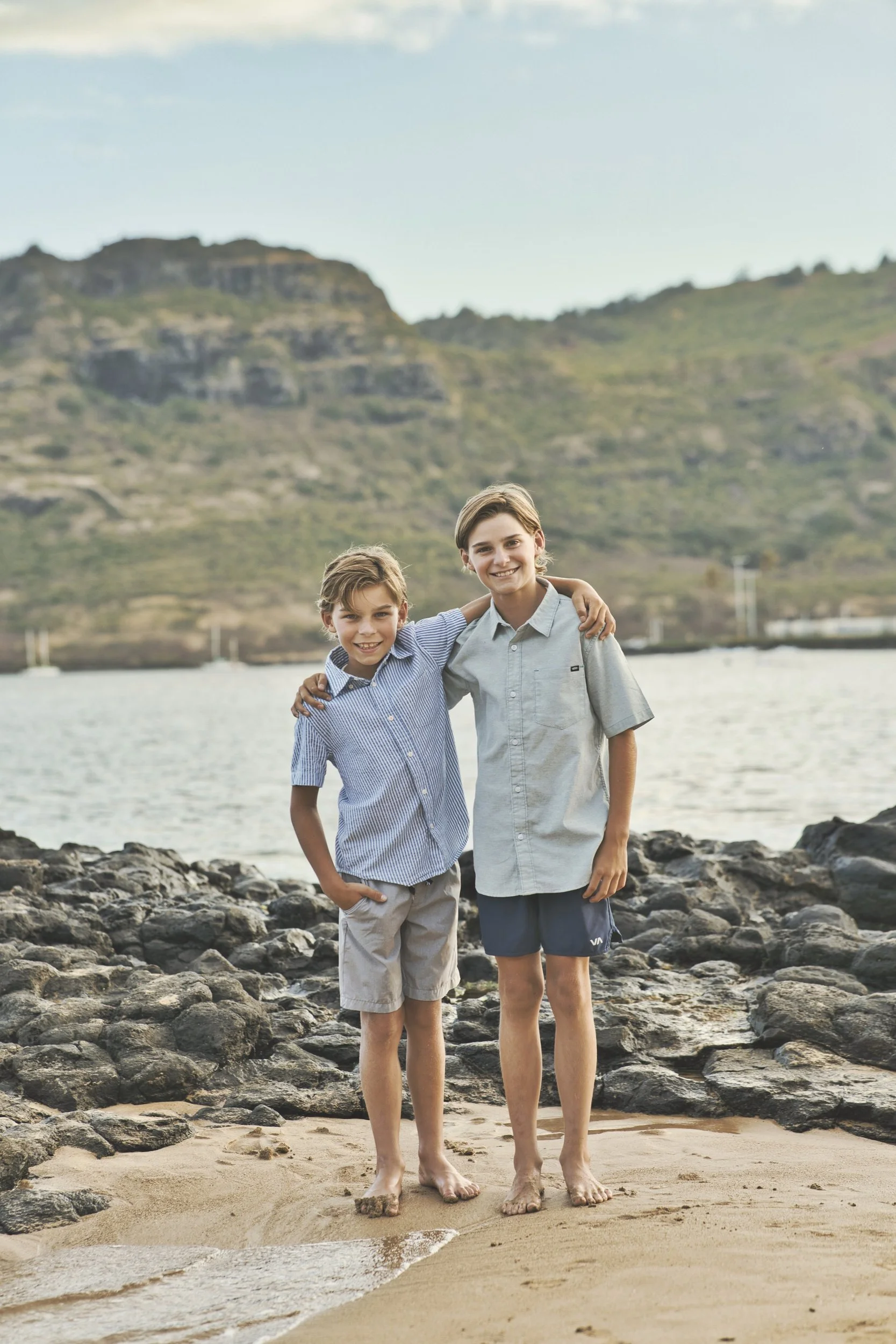 Two smiling brothers with arms around each other on the rocky shore of a Kauai bay.