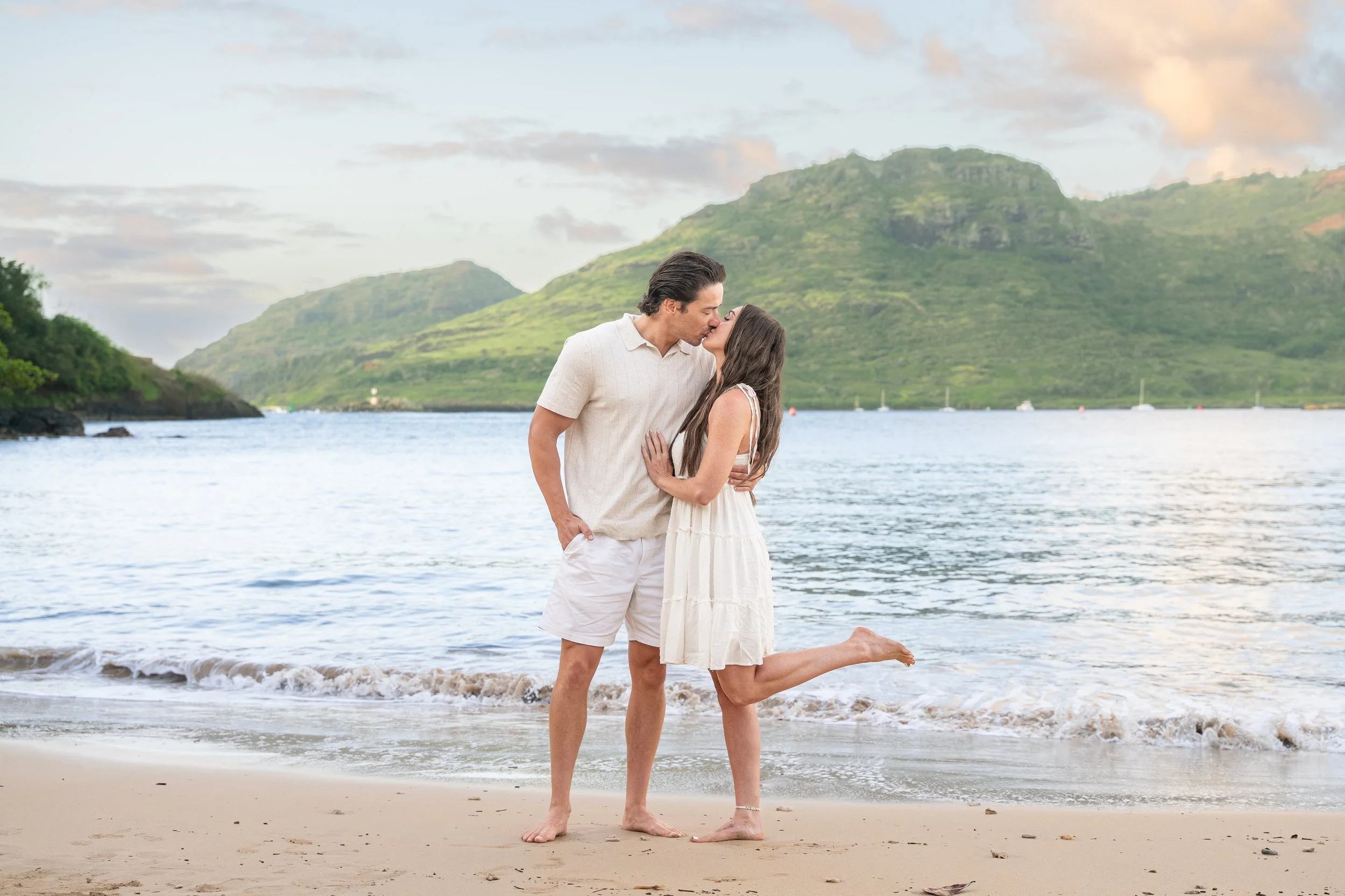 Couple kissing on a Kauai shoreline with the woman playfully kicking up her leg.
