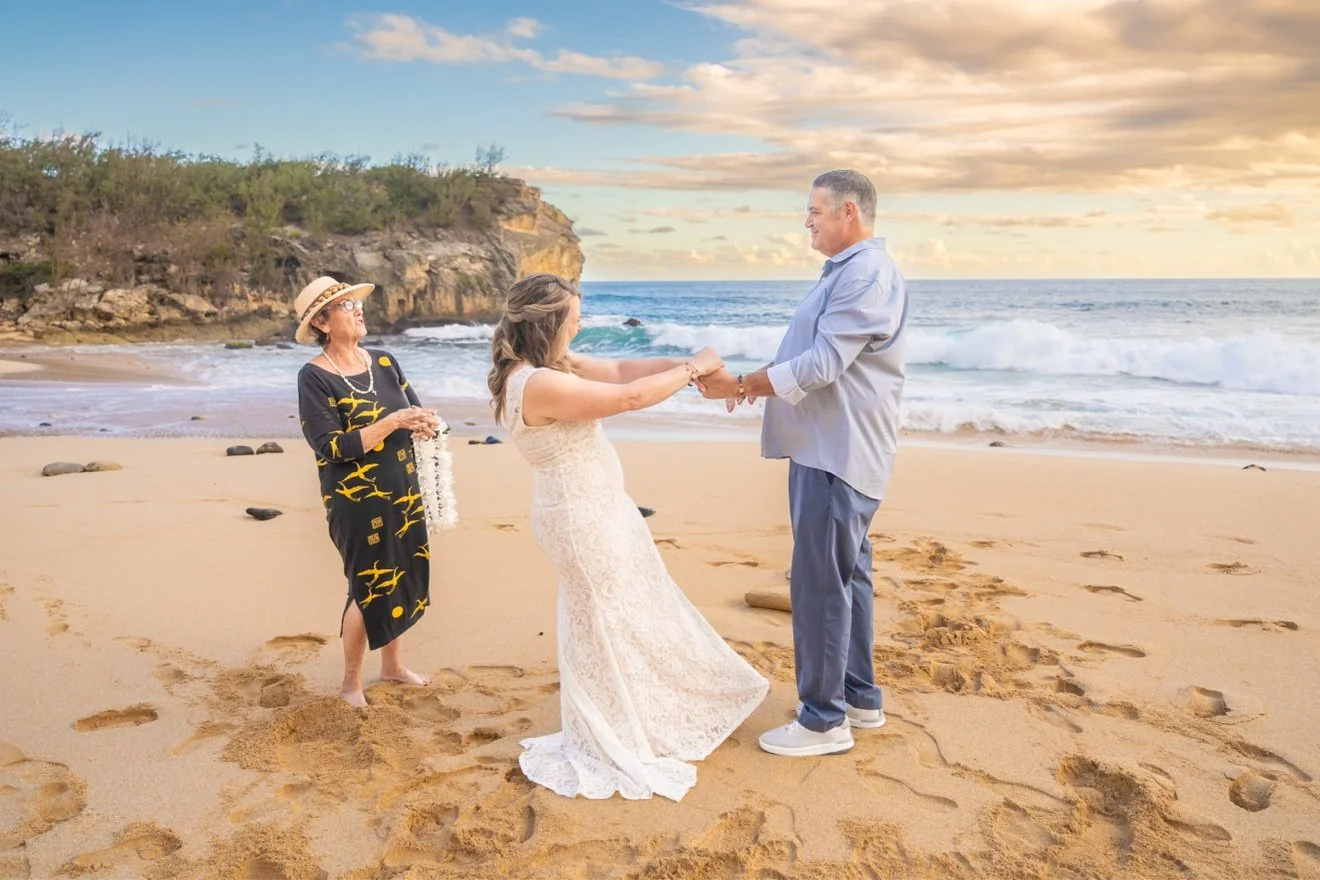 Bride and groom holding hands and facing each other during their beach wedding ceremony on Kauai, their officiant standing beside them with the ocean and cliffs behind.