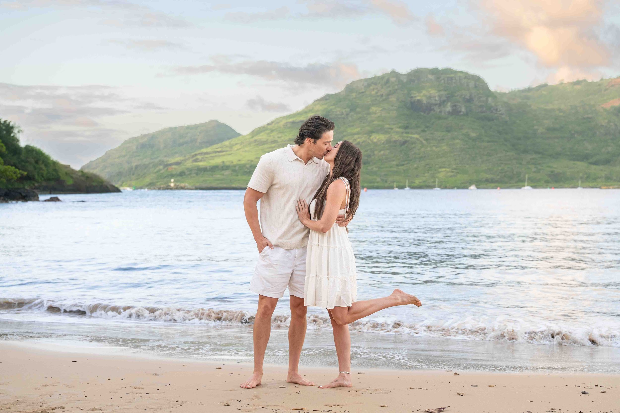 Couple kissing on a Kauai beach with green mountains behind them.