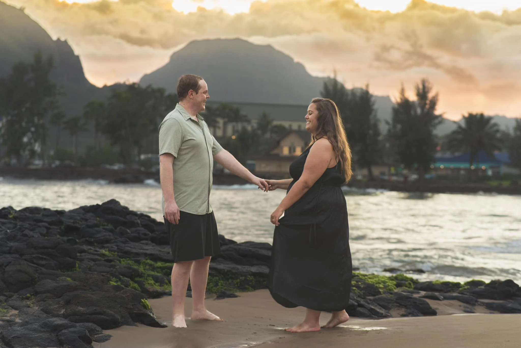 Couple holding hands and smiling at each other on a rocky Kauai shore at sunset.