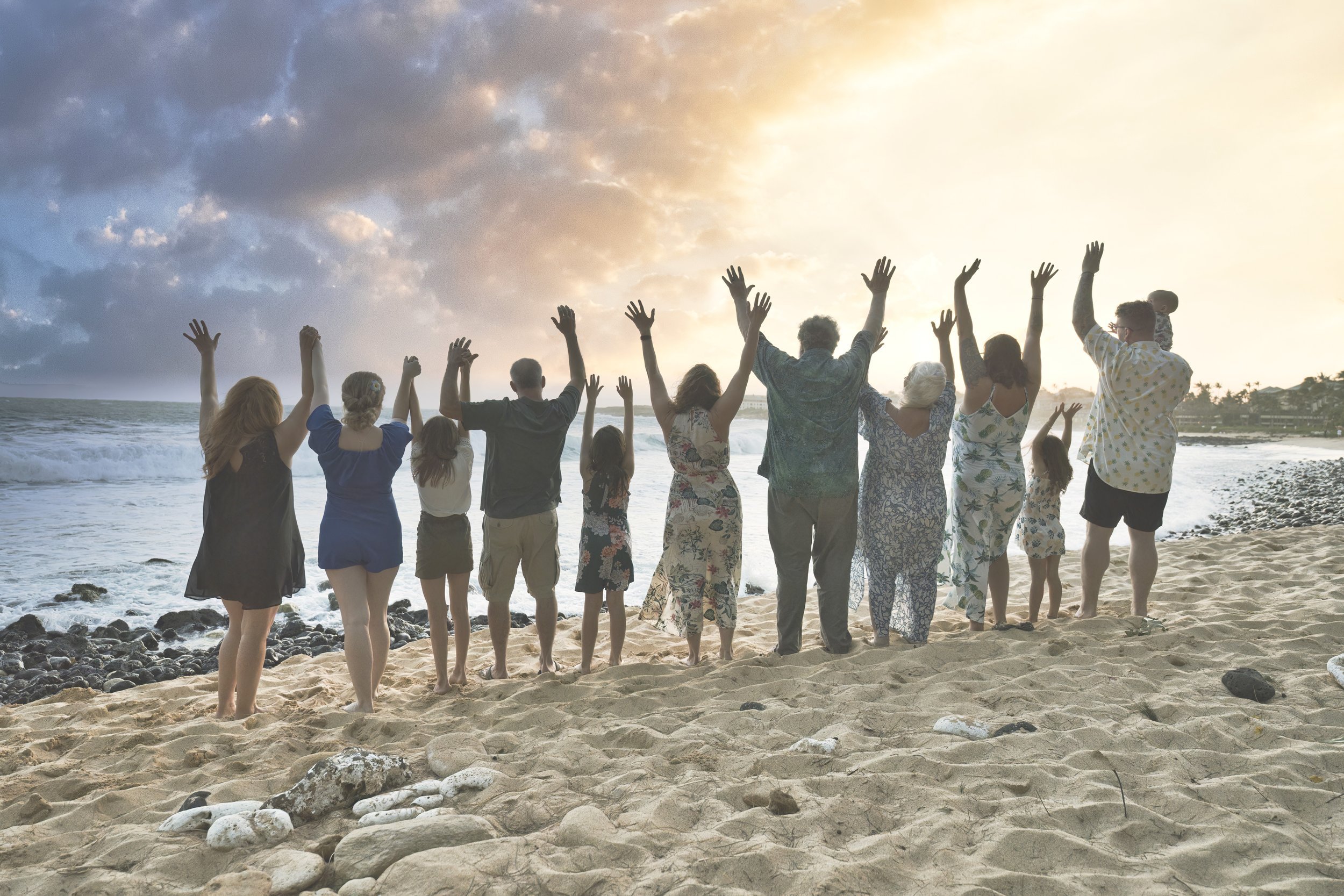 Large family standing with arms raised facing the sunset over the ocean on a Kauai beach.