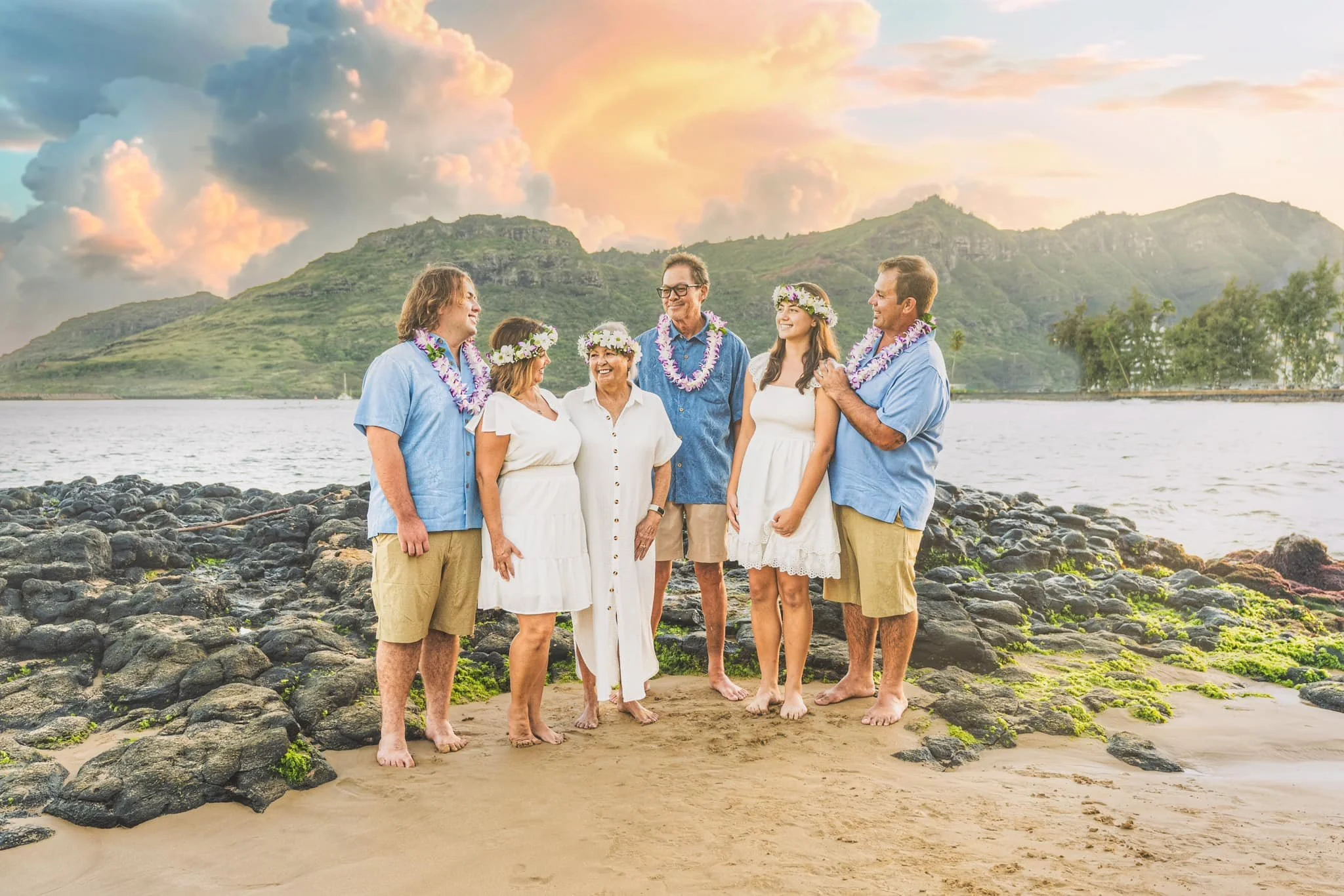 Extended family wearing flower leis poses on a rocky Kauai beach at sunset.