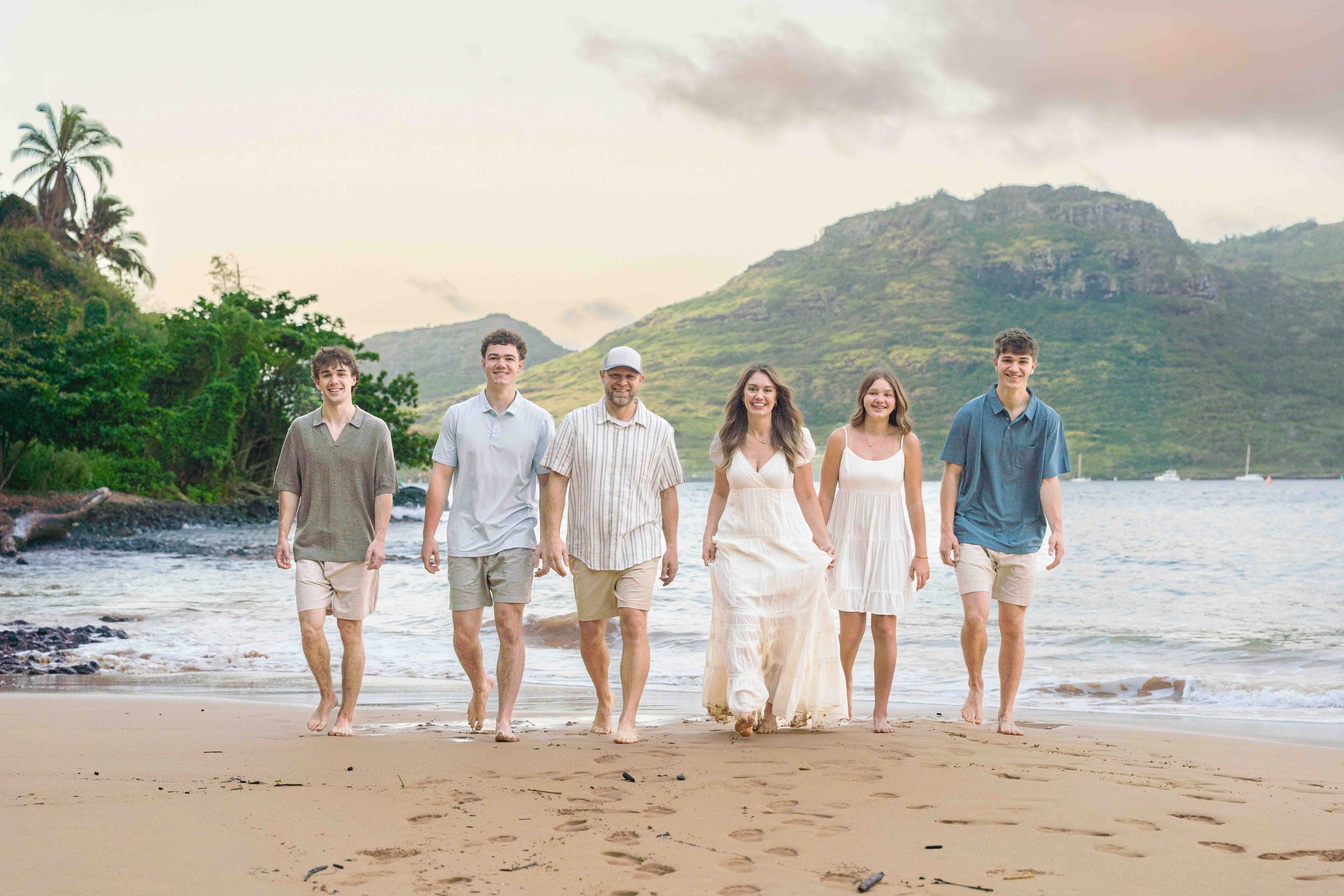 Family of six walking toward the camera on a Kauai beach with green mountains.