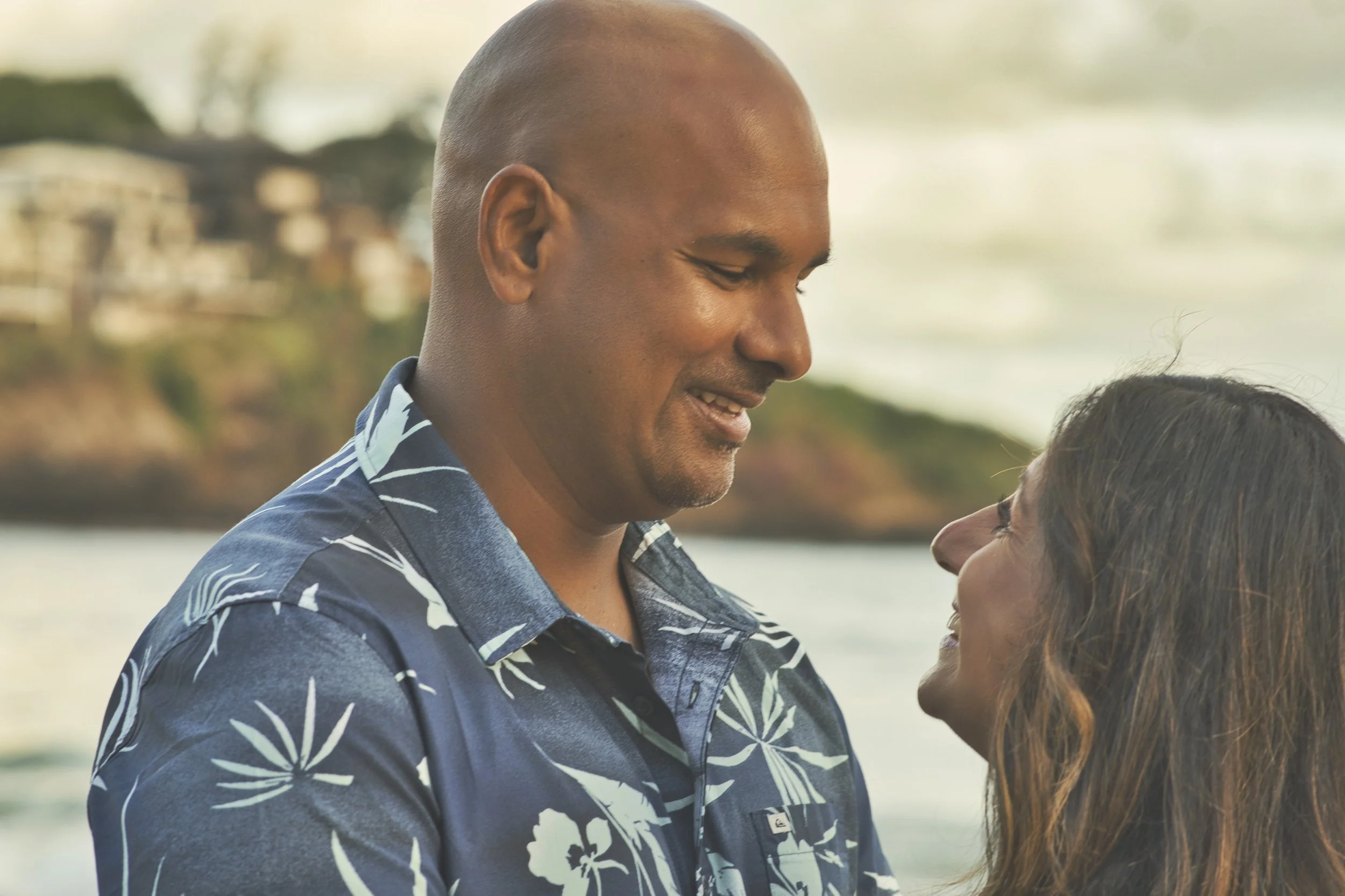 Couple smiling at each other on a Kauai beach with the coastline behind them.