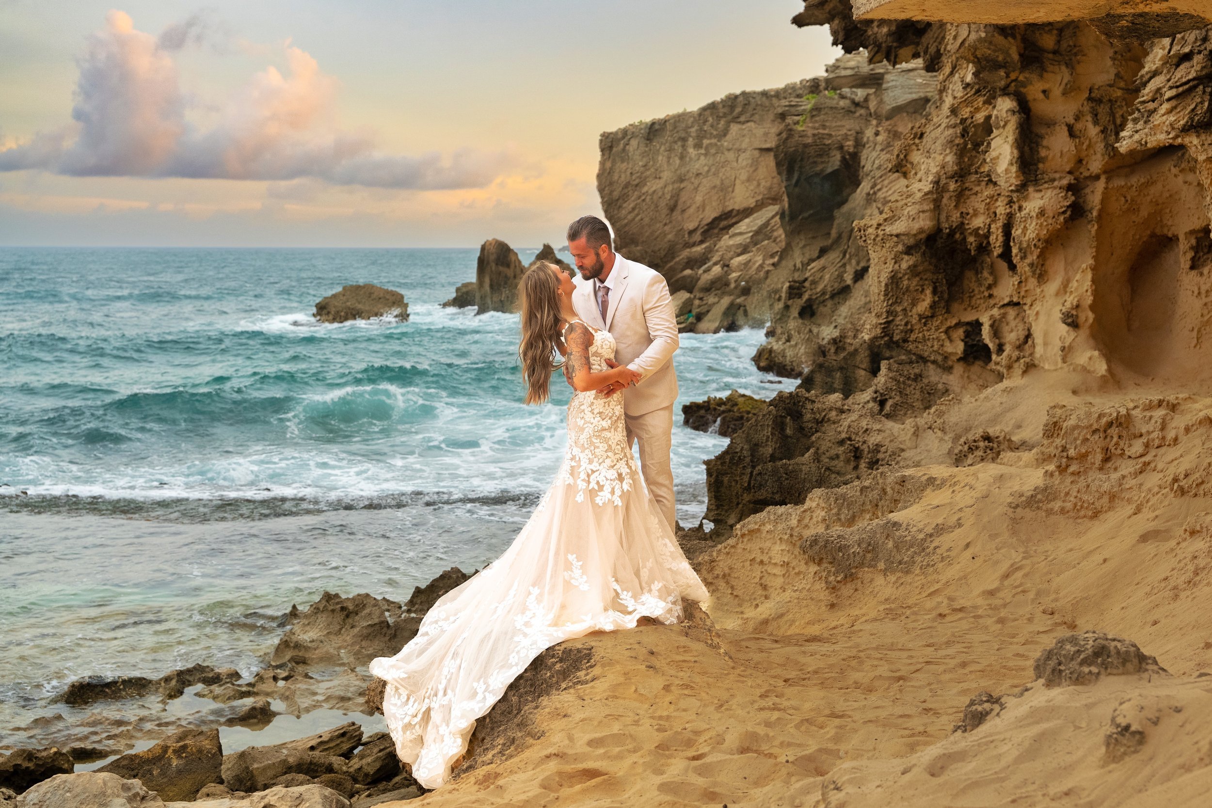 Newlywed couple kissing on a rocky Kauai shoreline with waves crashing nearby.