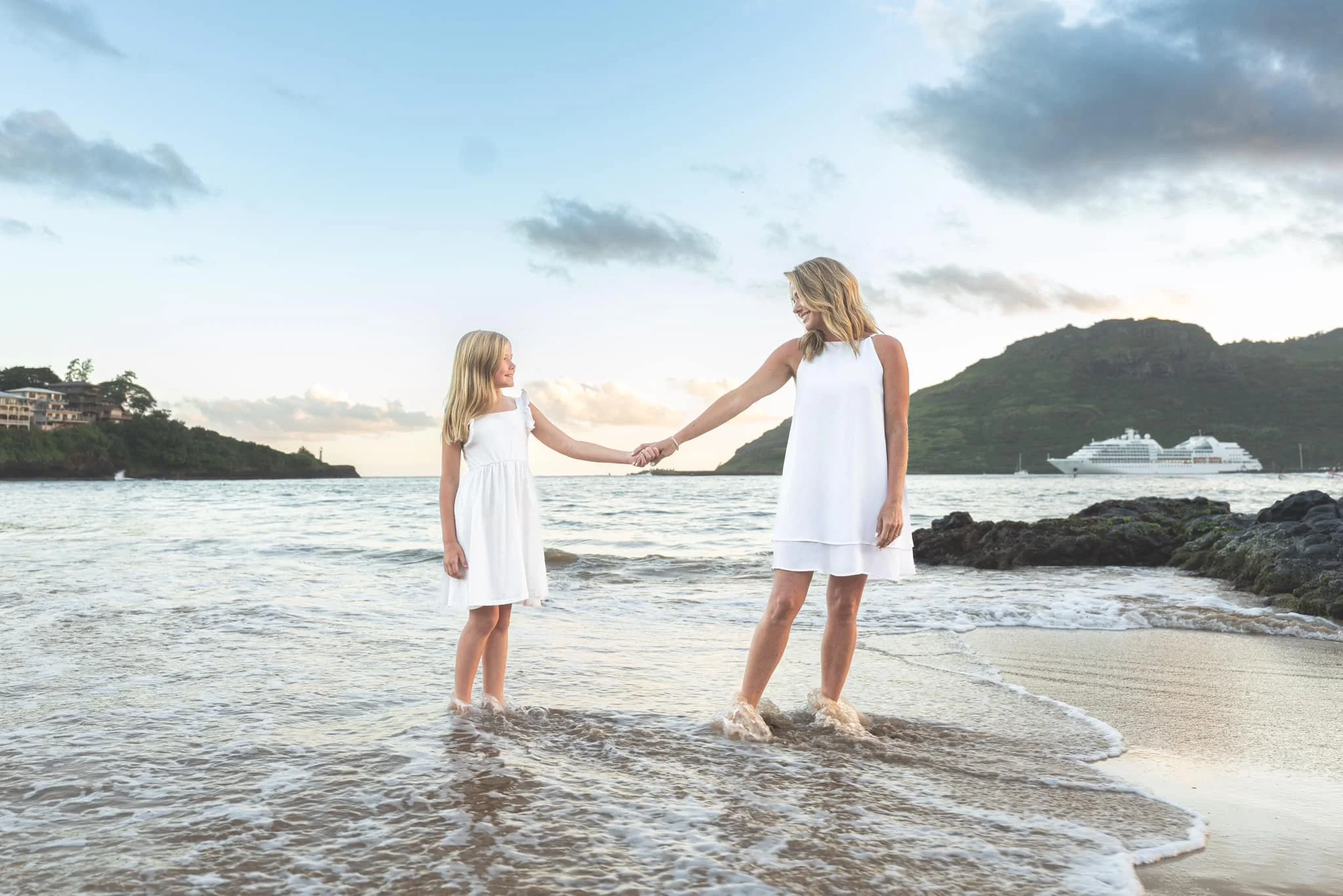 Mother and daughter in white dresses holding hands while wading in the Kauai surf.