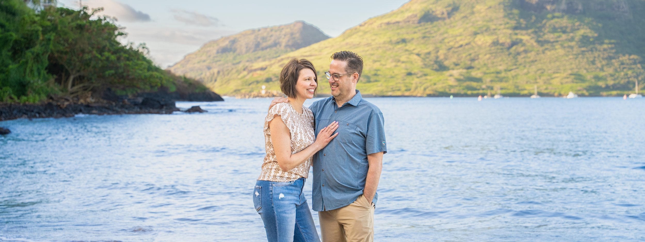 Smiling couple embracing by a calm Kauai bay with green mountains behind them.
