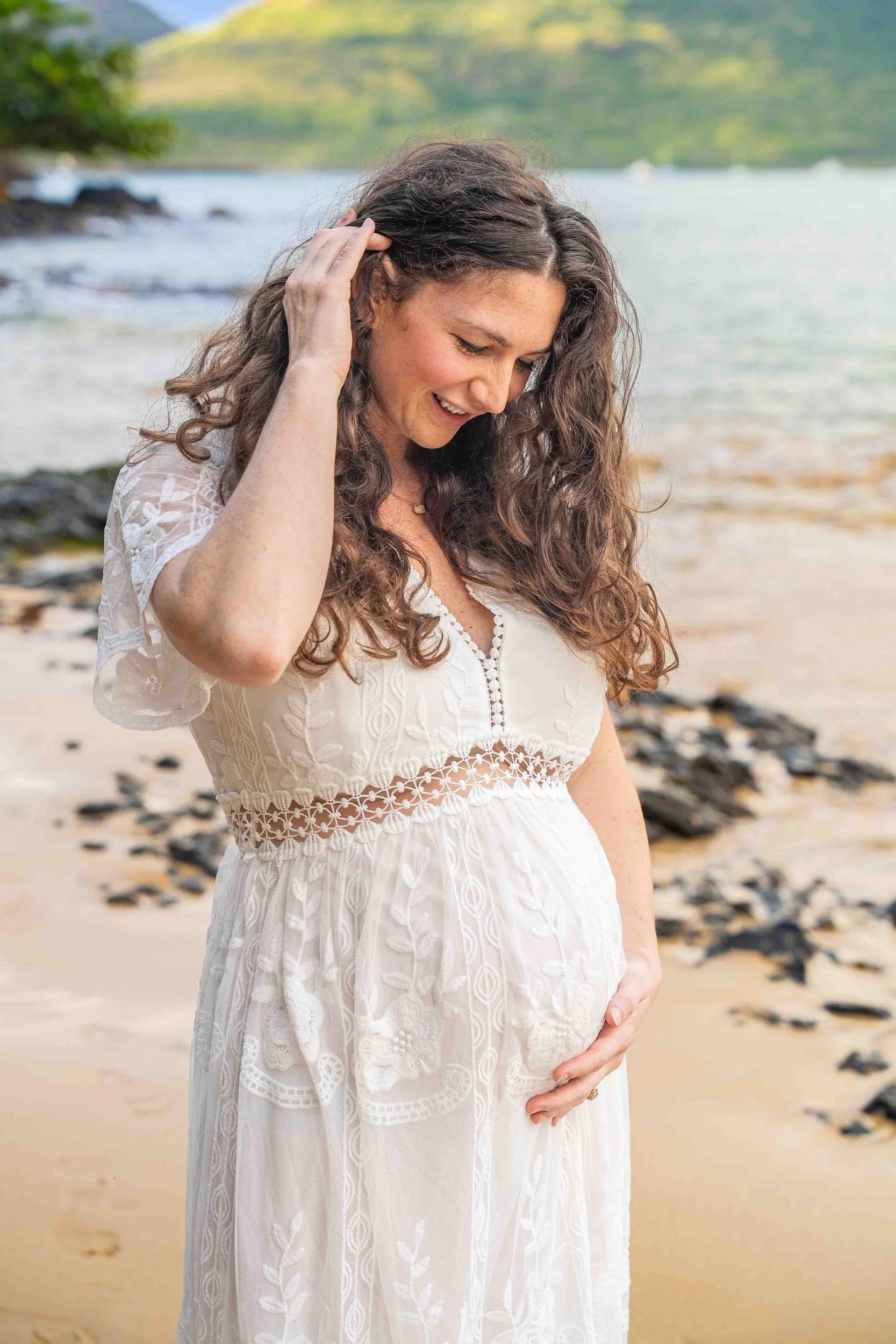 Expecting mom in a white lace dress looking down at her belly on a Kauai beach.