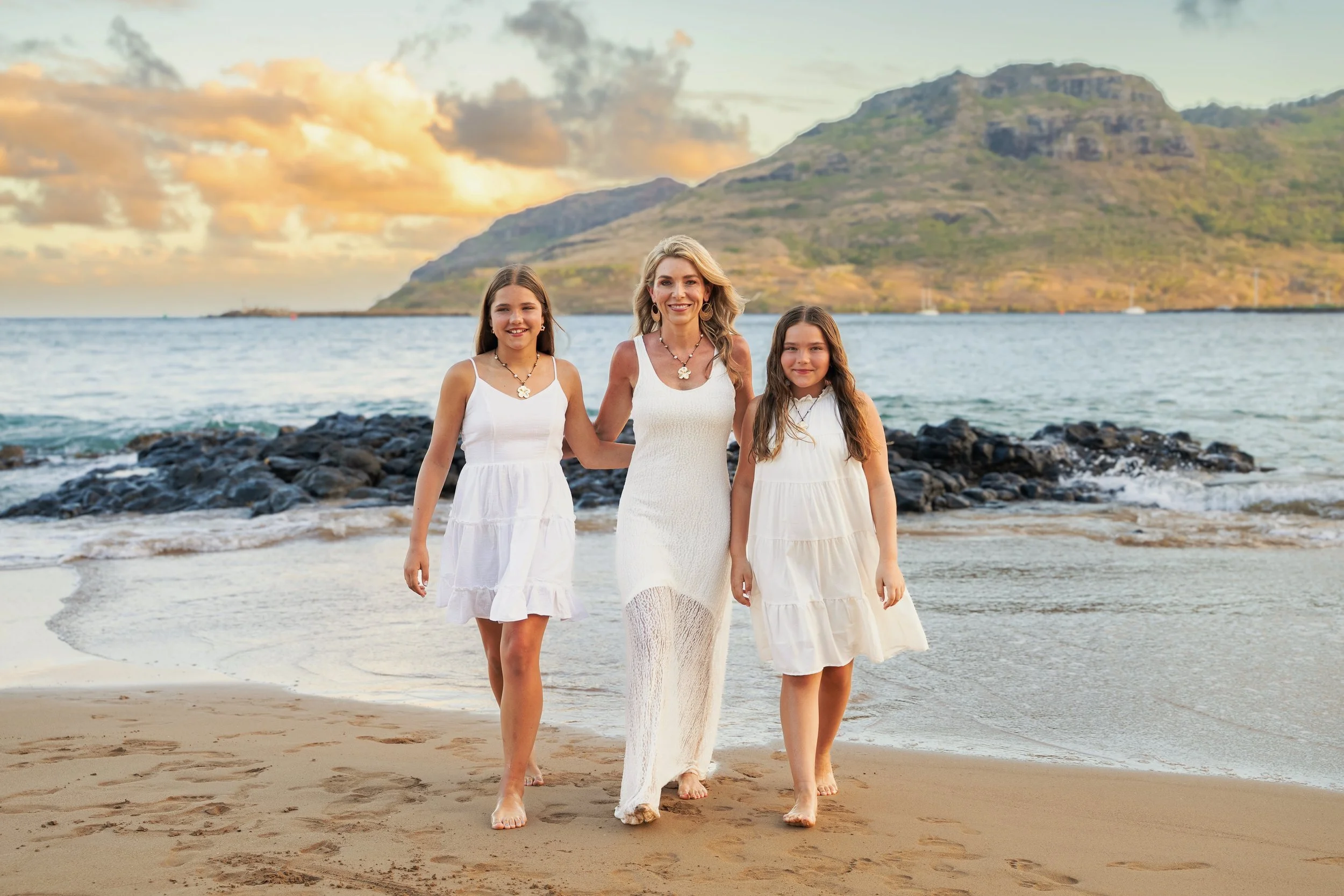 Mom walking on a Kauai beach with her two daughters in white dresses at sunset.