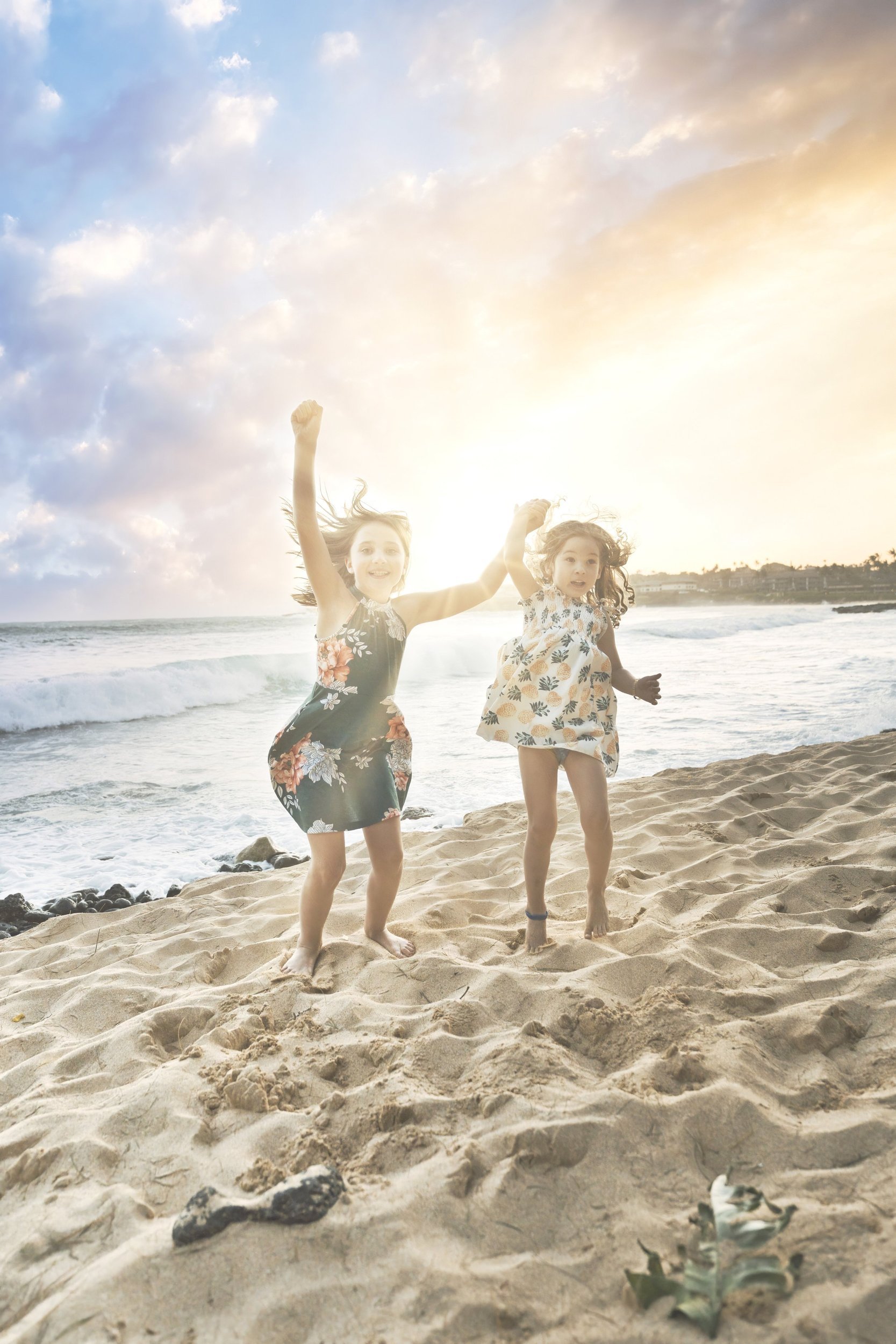 Two young girls dancing joyfully on a sandy Kauai beach at sunset.