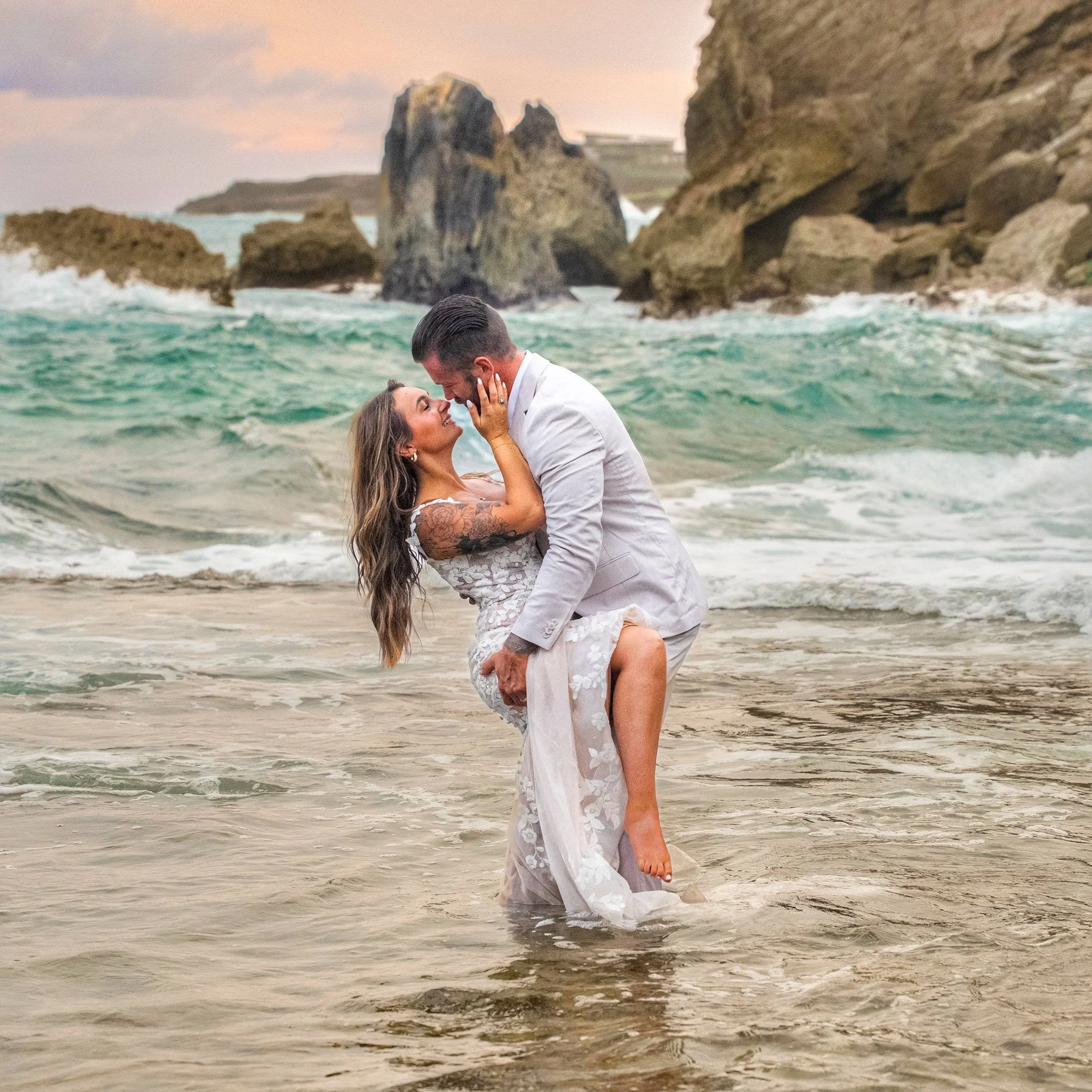 Groom lifting his bride for a kiss while standing in the ocean surf on a Kauai beach at sunset.