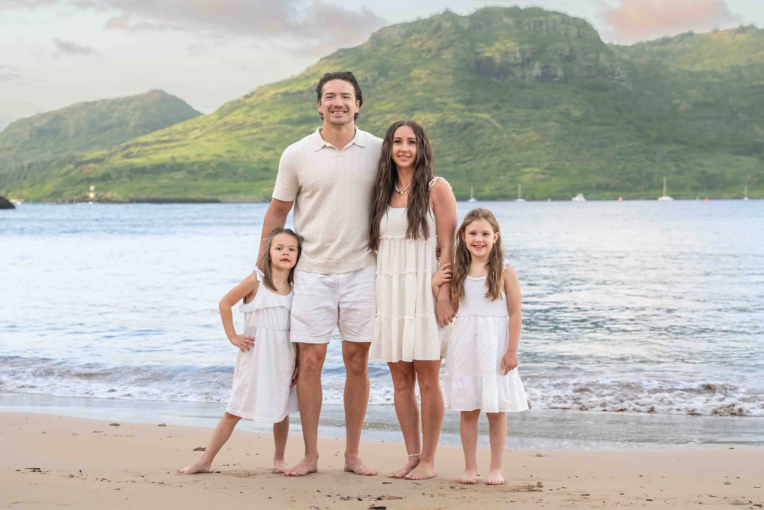 Family of four in white outfits posing on a Kauai beach with a mountain behind them.