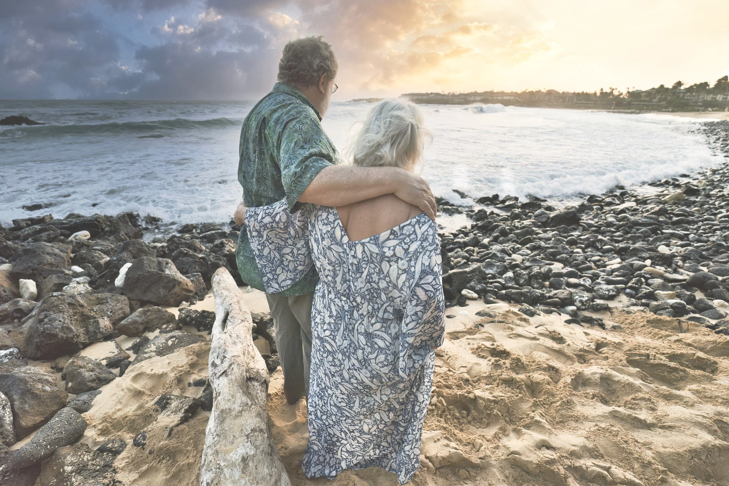 Older couple embracing while watching the sunset over a rocky Kauai beach.