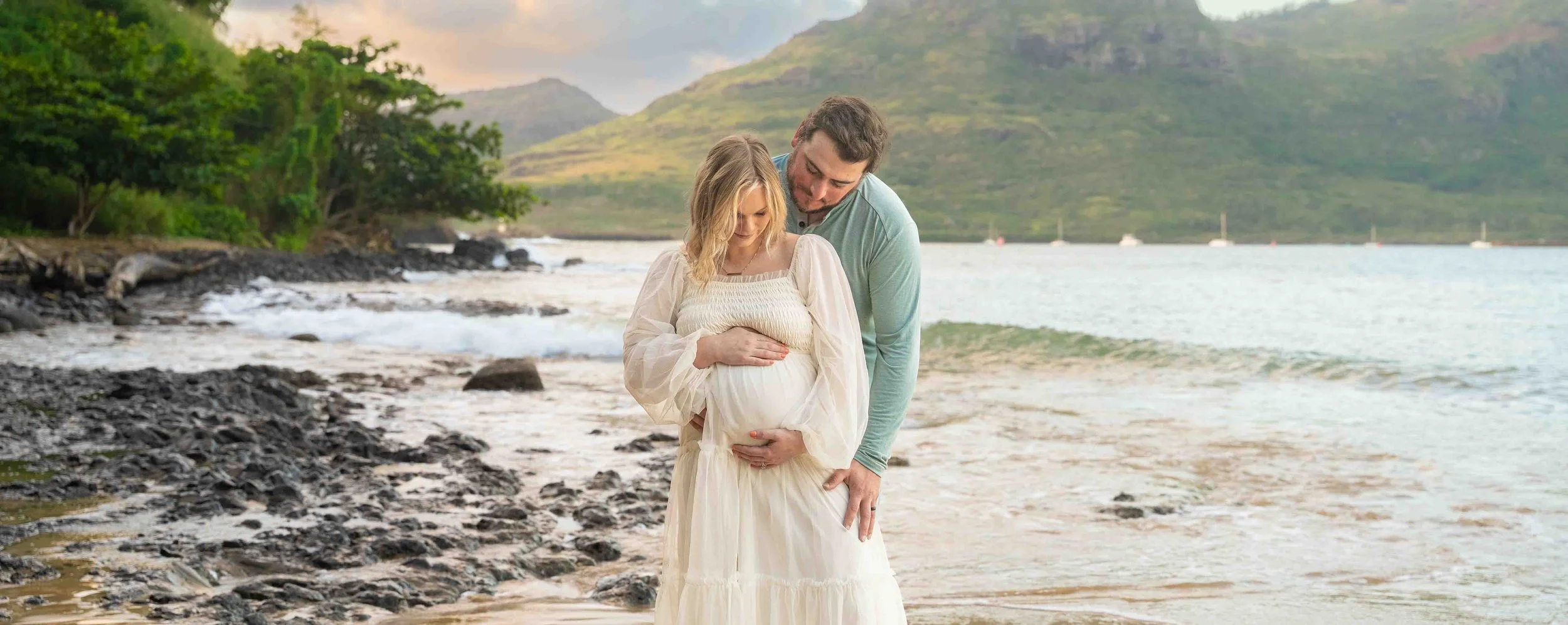Pregnant woman in a white dress with her partner on a rocky Kauai shore.