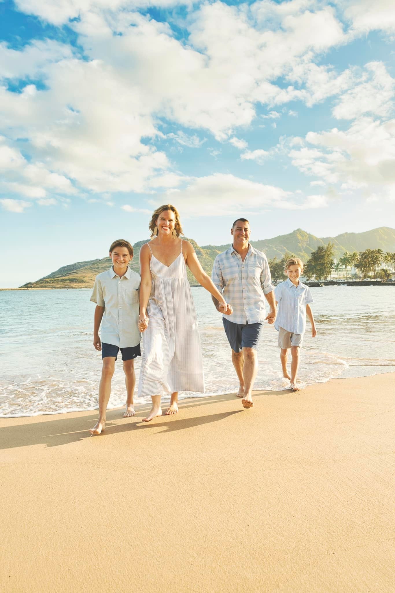 Family of four holding hands and walking along a sandy Kauai beach with palms and mountains behind.