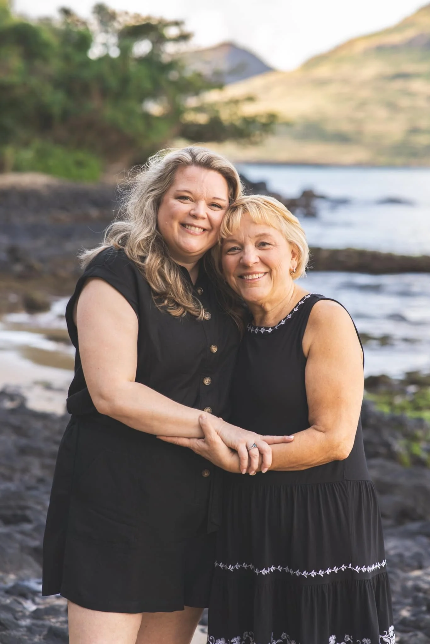 Mother and daughter embrace on a rocky Kauai shoreline, both wearing black dresses.