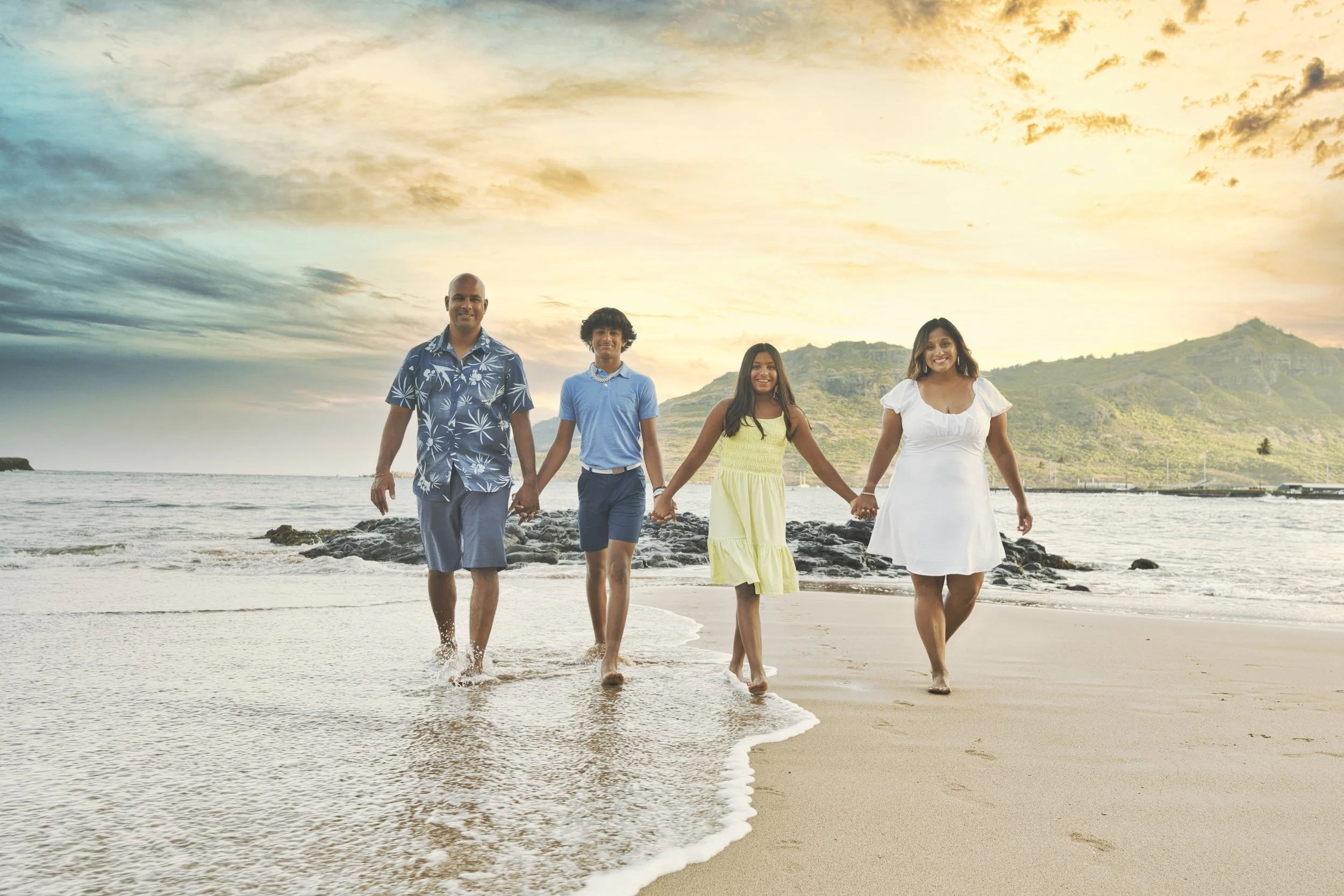 The family walks hand-in-hand through golden surf as the sun sets behind the Kauai mountains.