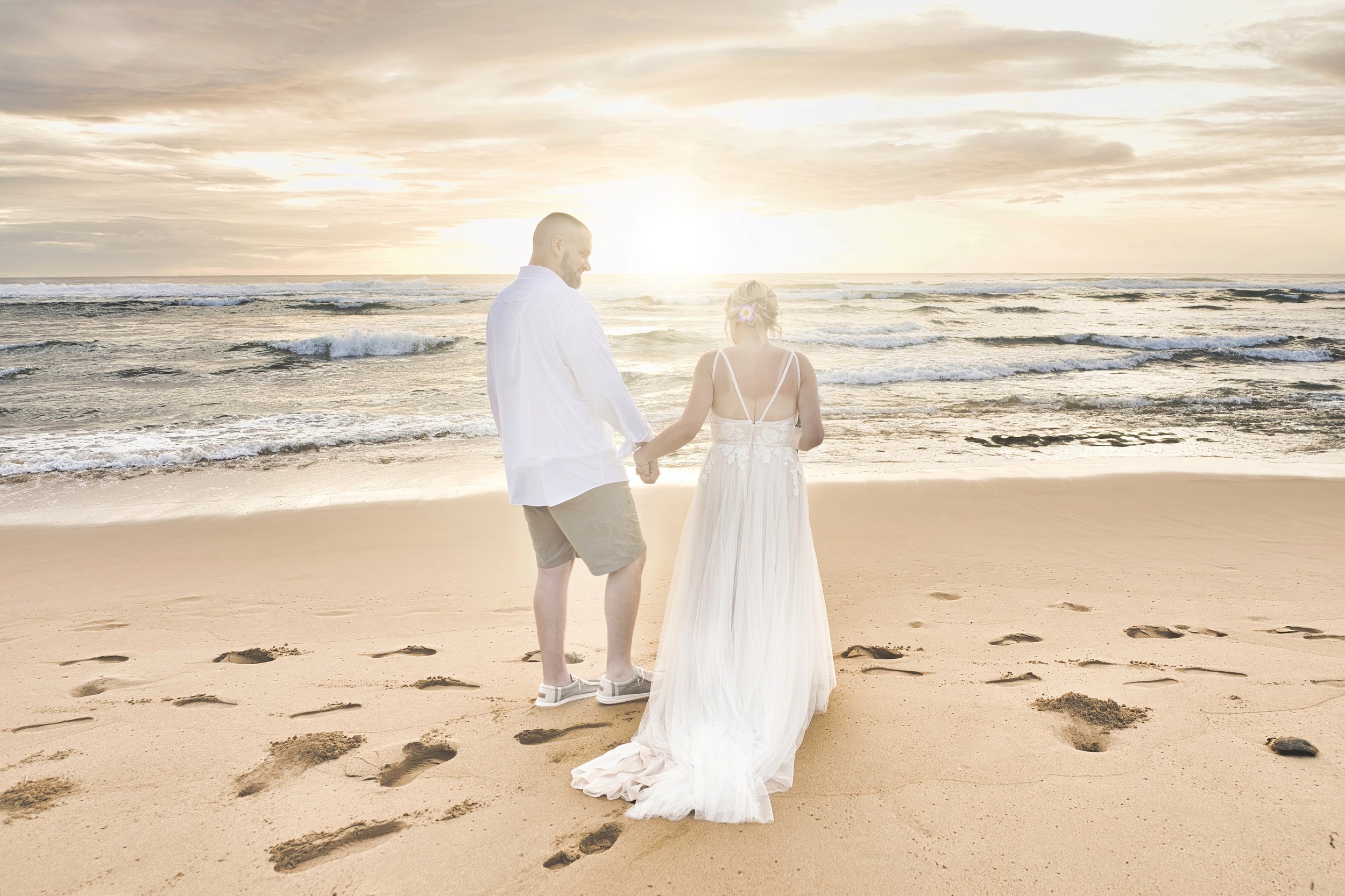 Newlywed couple walking hand in hand toward the ocean on a Kauai beach at sunset.