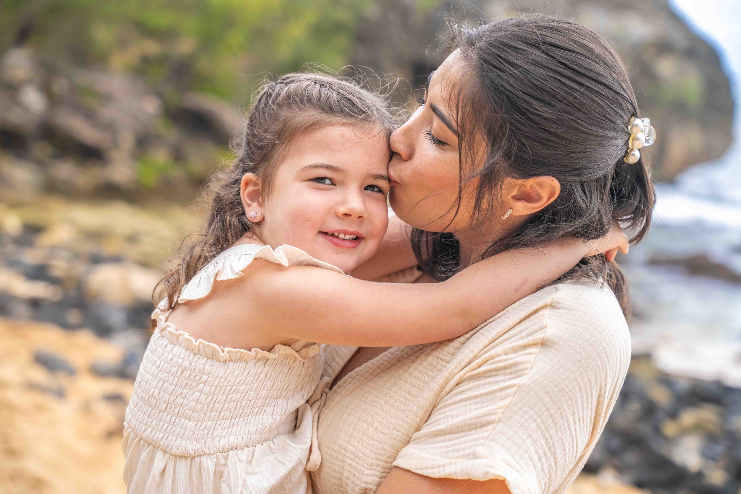 Mom kissing her young daughter on the forehead at a Kauai beach.