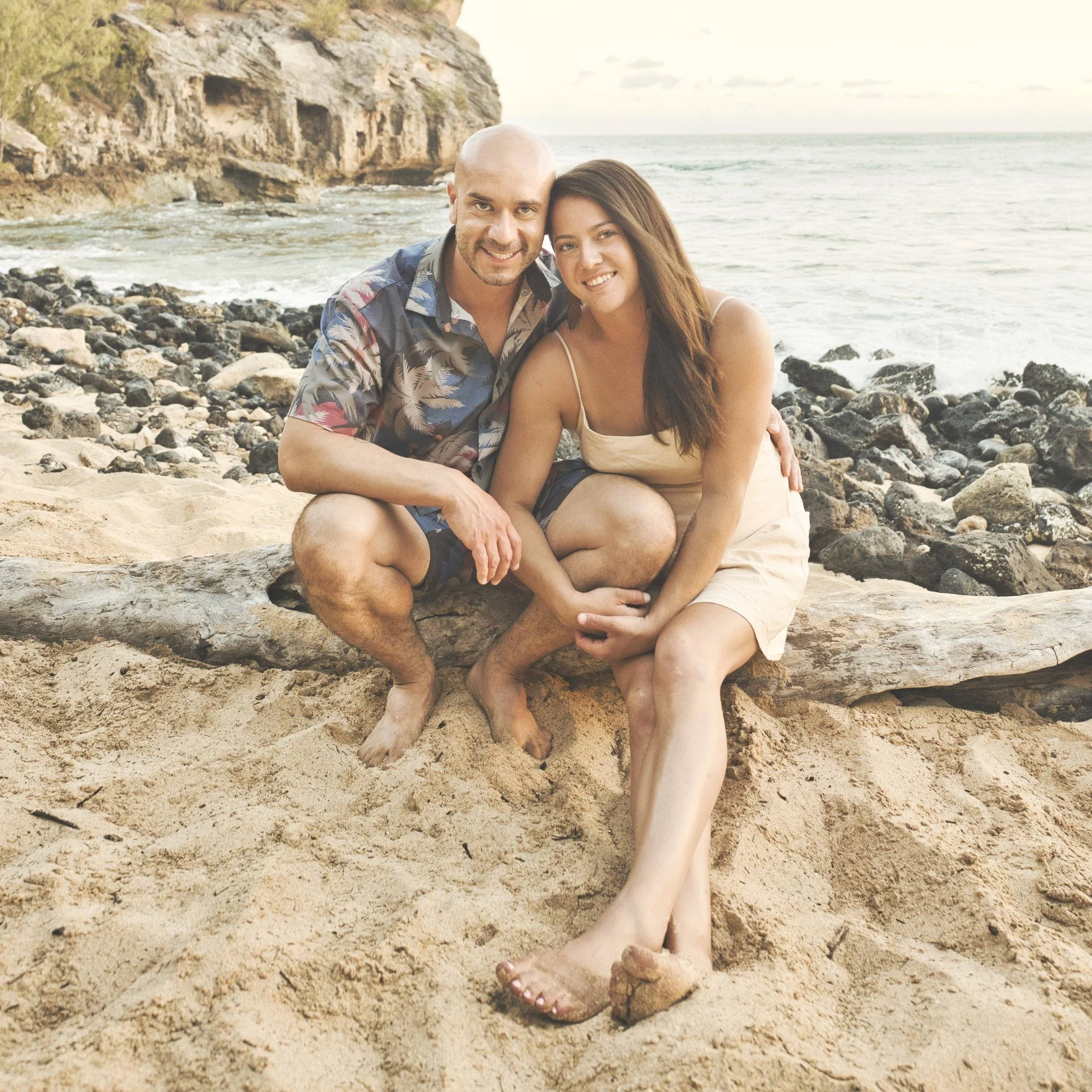 Smiling couple sitting on driftwood on the beach at Shipwreck Beach, Kauai.
