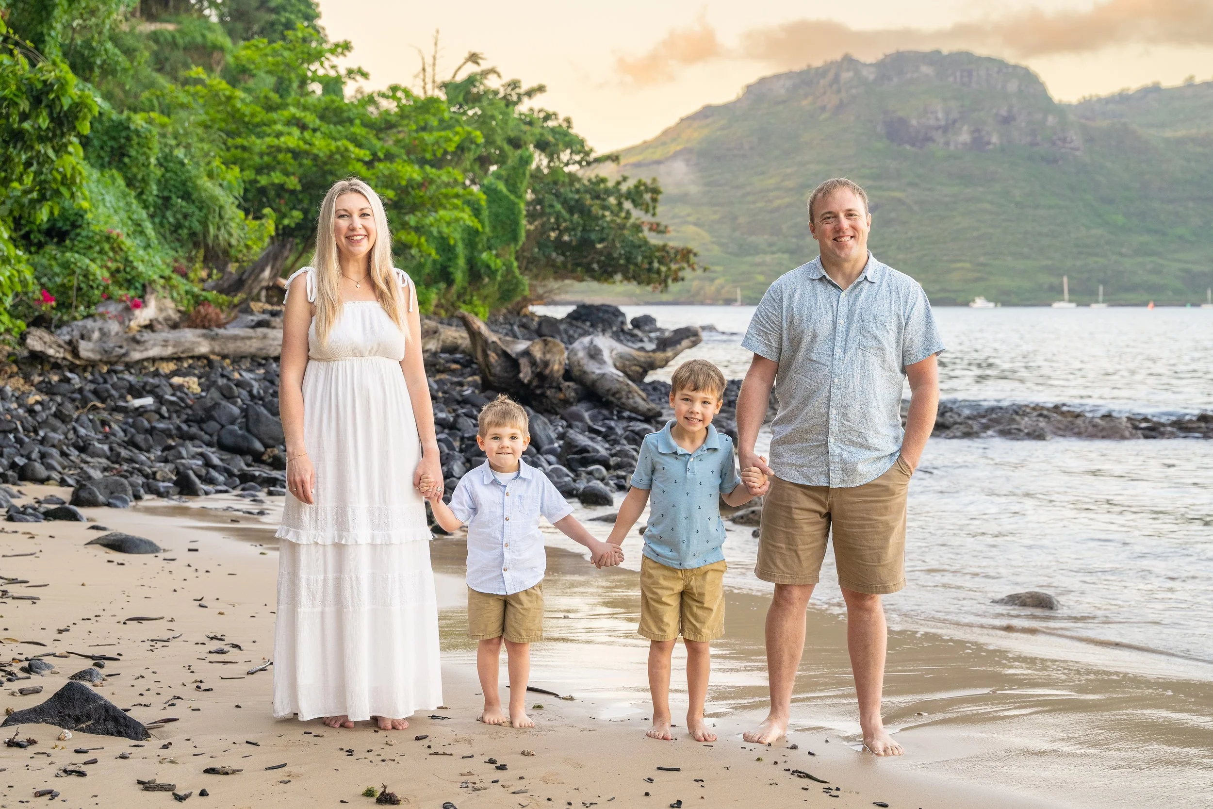 Family of four holding hands on a rocky Kauai beach with a mountain backdrop.