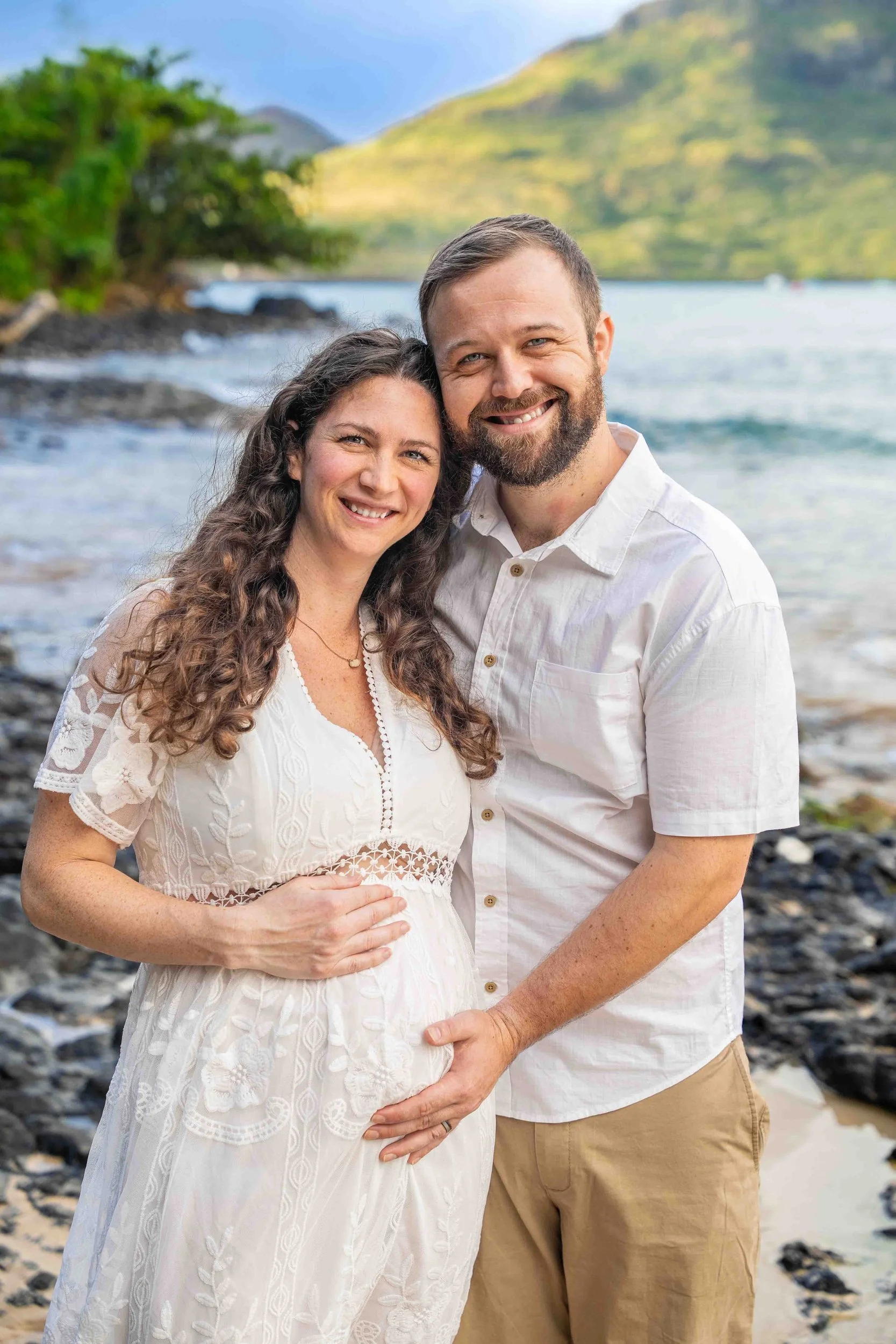 Smiling expecting couple in white on a rocky Kauai shore.