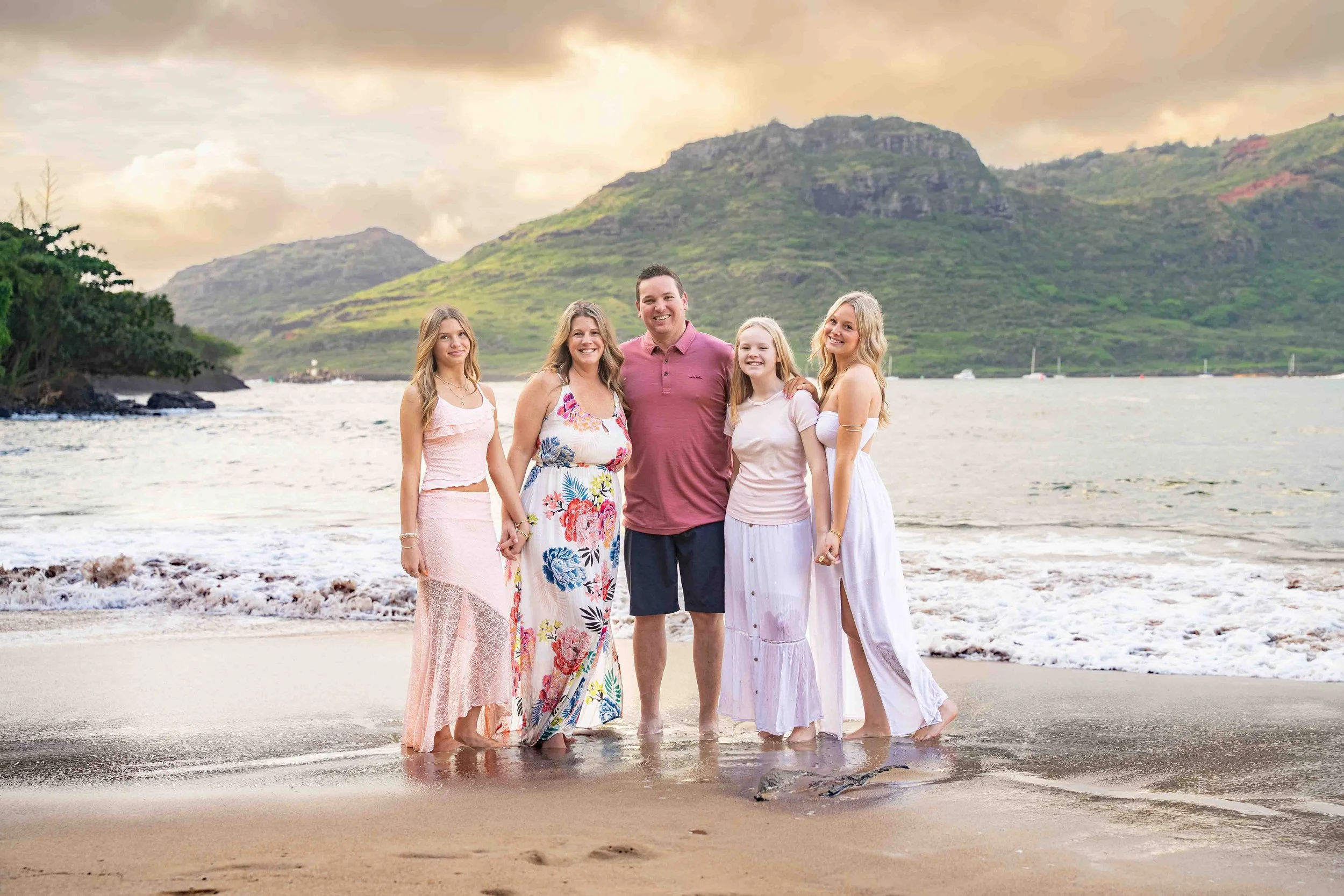 Family of five standing in the surf on a Kauai beach with a mountain behind them.