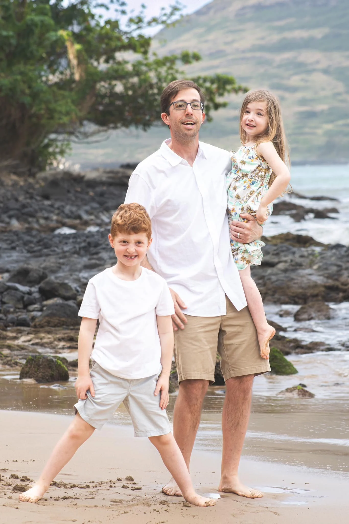 Dad holding his little daughter with his son beside them on a rocky Kauai shore.