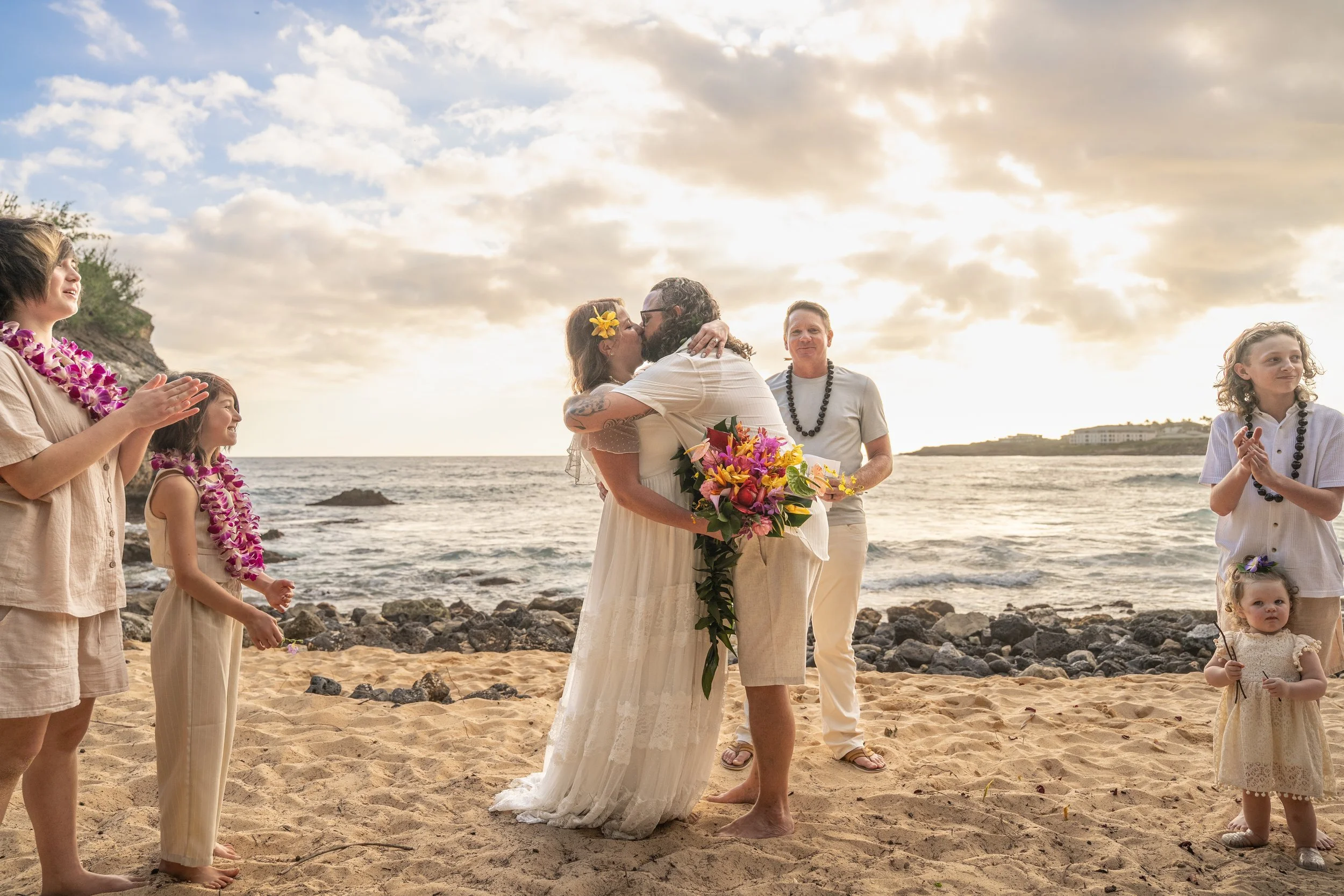 Bride and groom sharing their first kiss as family applauds on a Kauai beach.