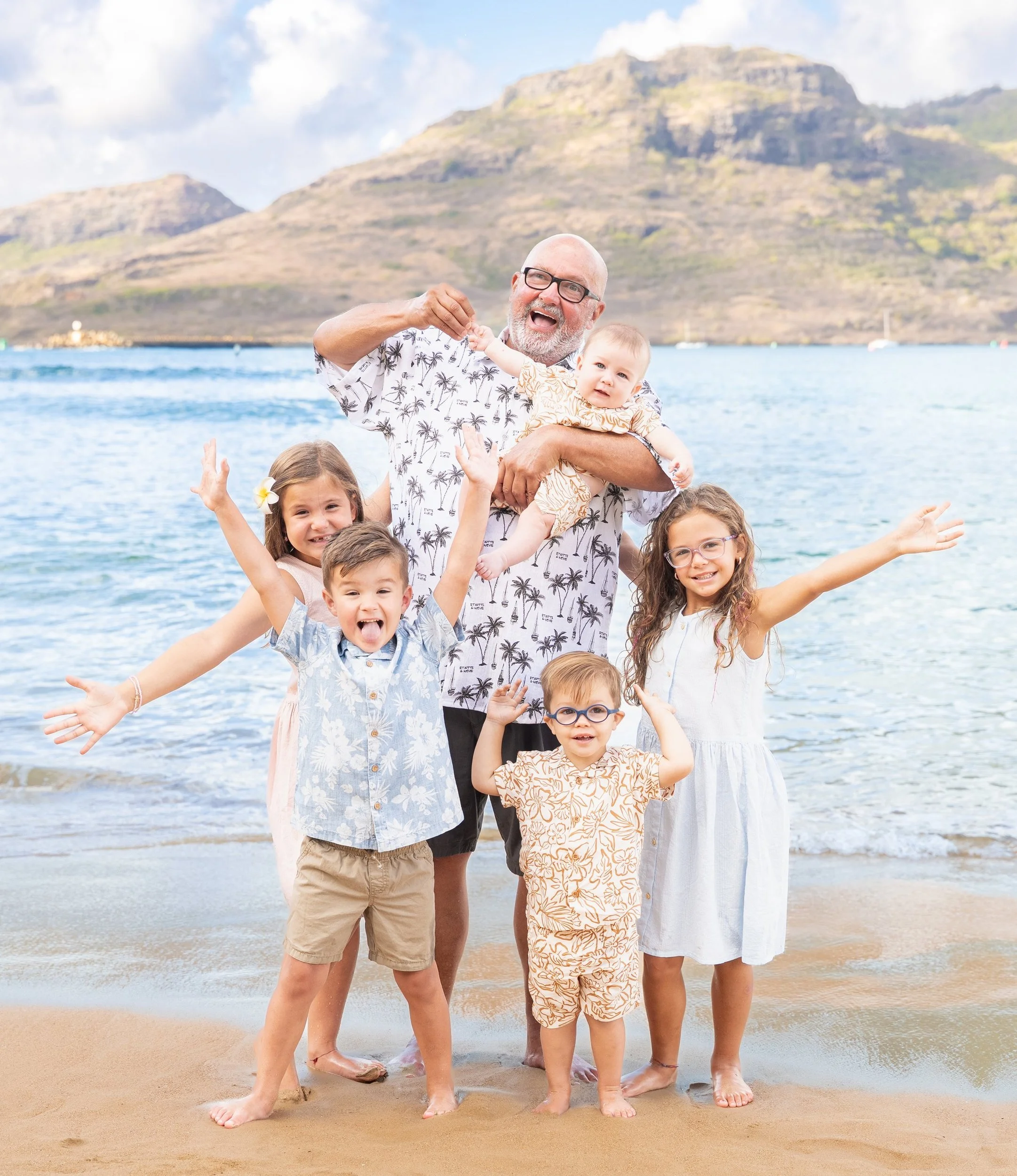 Grandpa holding a baby while four grandkids cheer with raised arms on a Kauai beach.