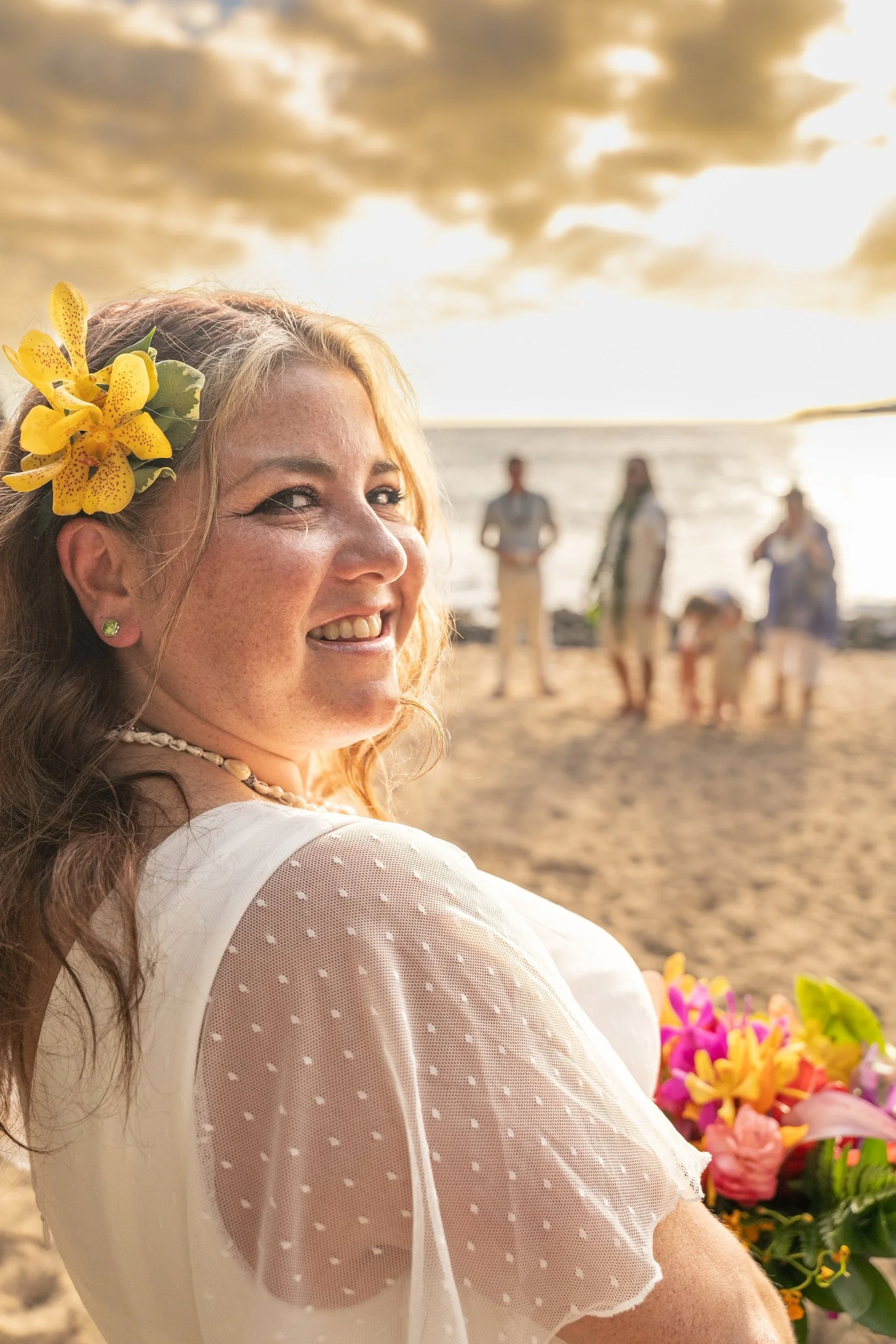 Smiling bride with an orchid in her hair holding a tropical bouquet on a Kauai beach.
