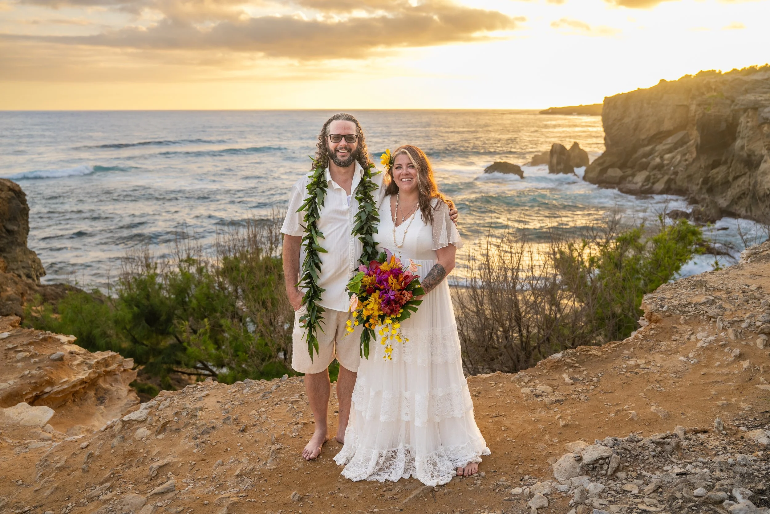 Newlywed couple posing on a Kauai cliffside at sunset, the bride holding a tropical bouquet.